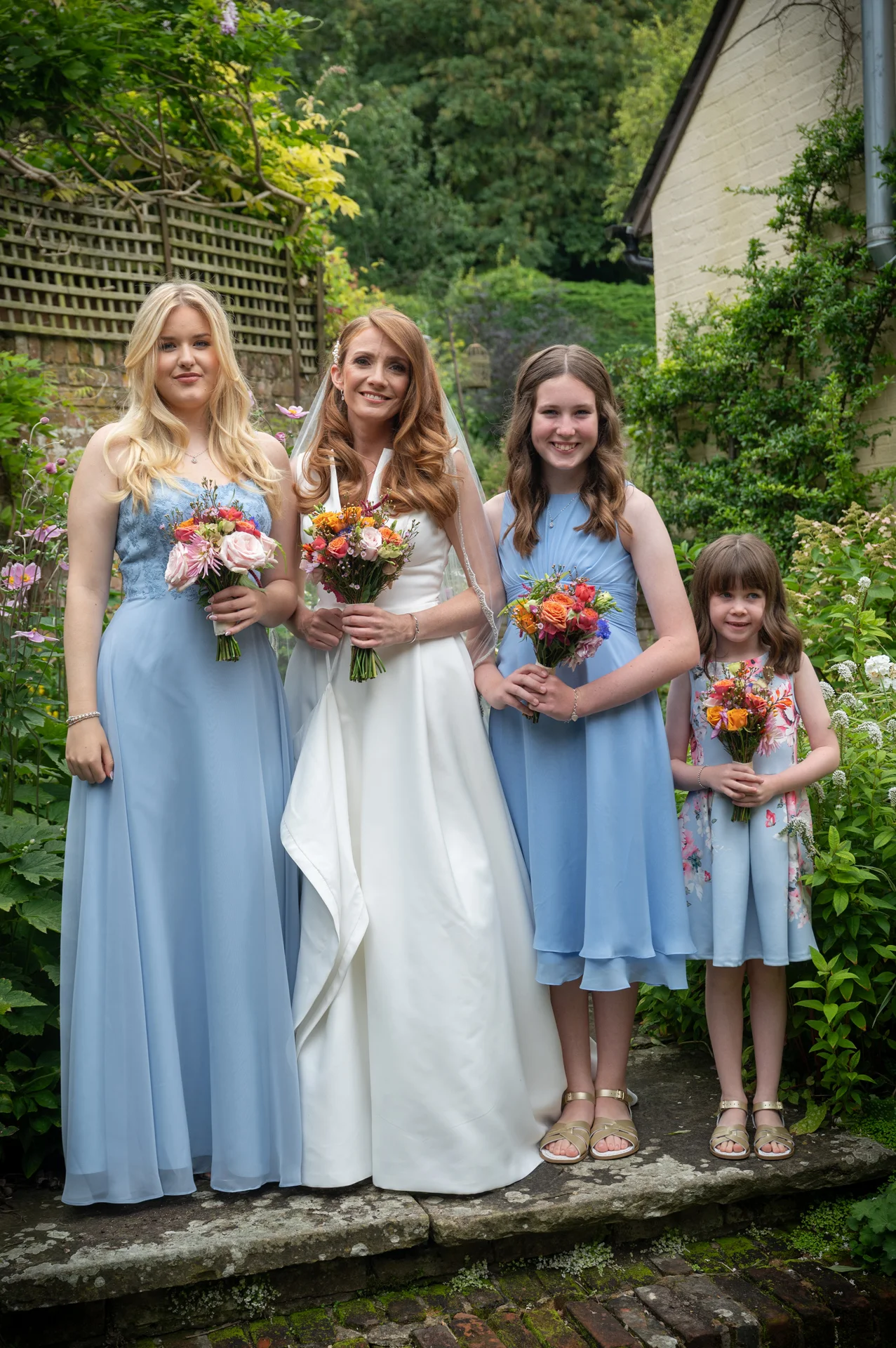 A smiling bride in a white a-line gown stands in a lush garden alongside two bridesmaids in blue dresses and a young flower girl, each holding a vibrant floral bouquet.