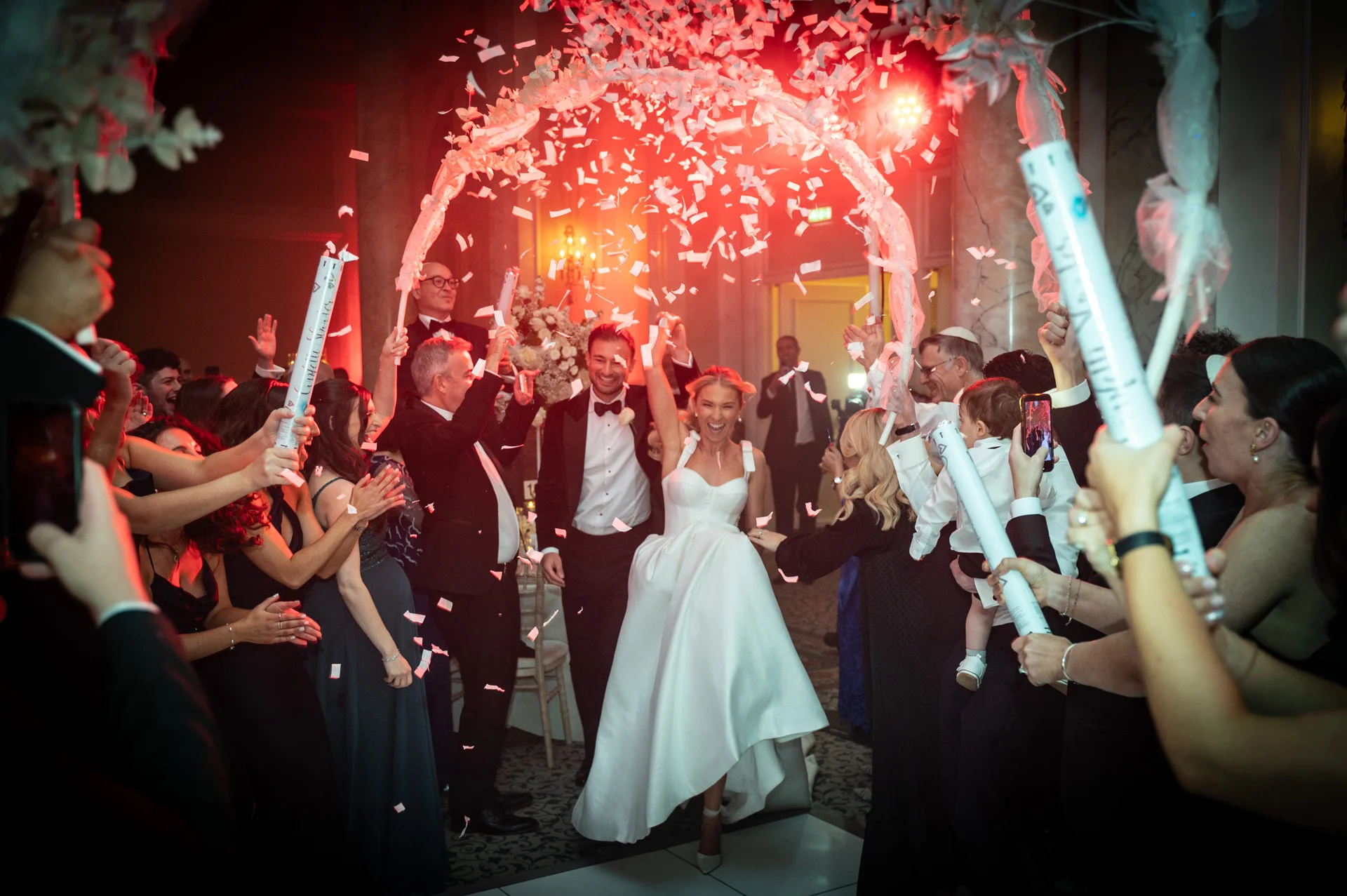 The joyful newlyweds make their grand entrance through a floral archway amidst a flurry of falling confetti and cheering guests.