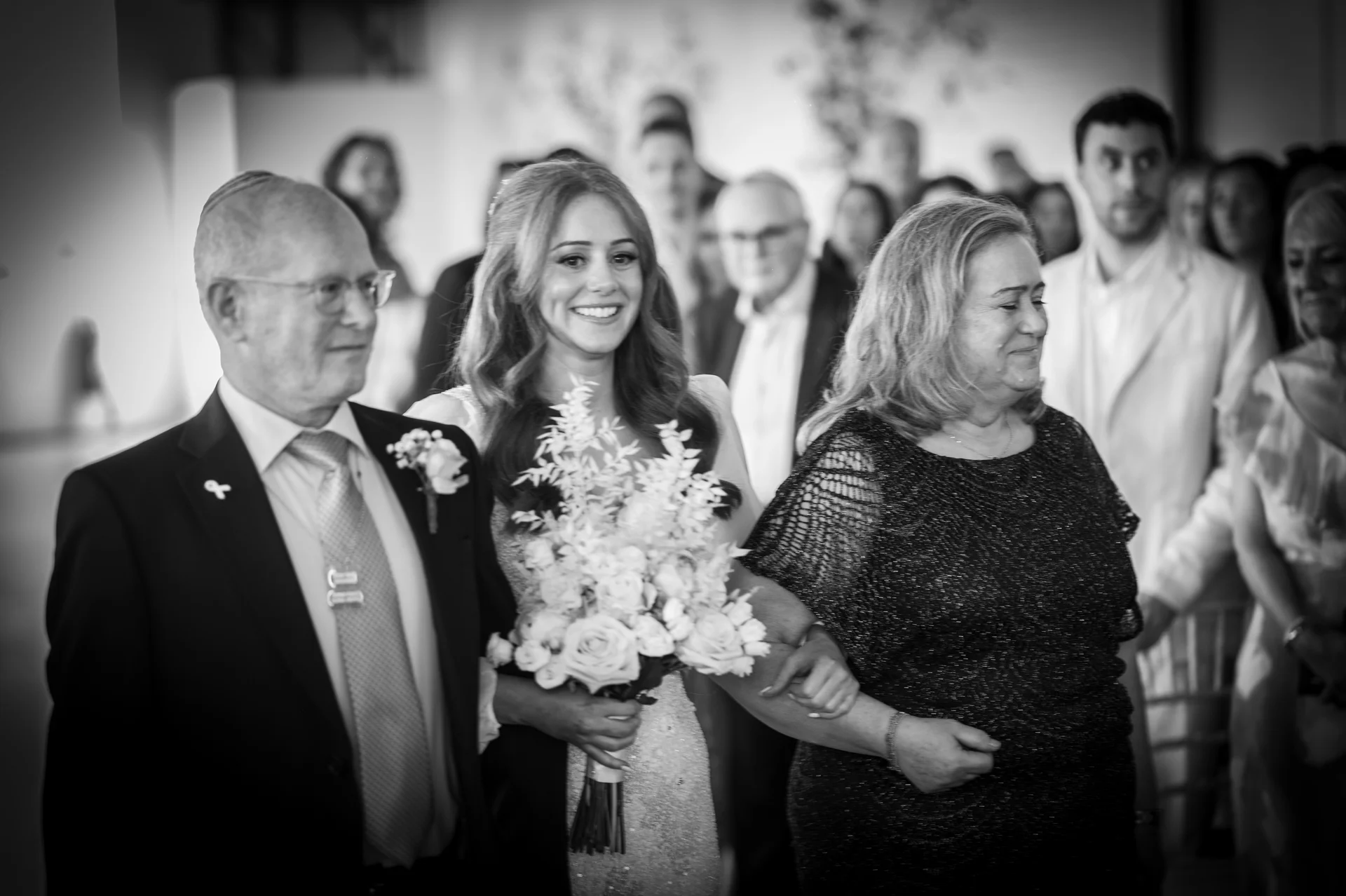 A joyful bride walking down the aisle arm in arm with her parents, holding a bouquet of roses during the wedding ceremony.