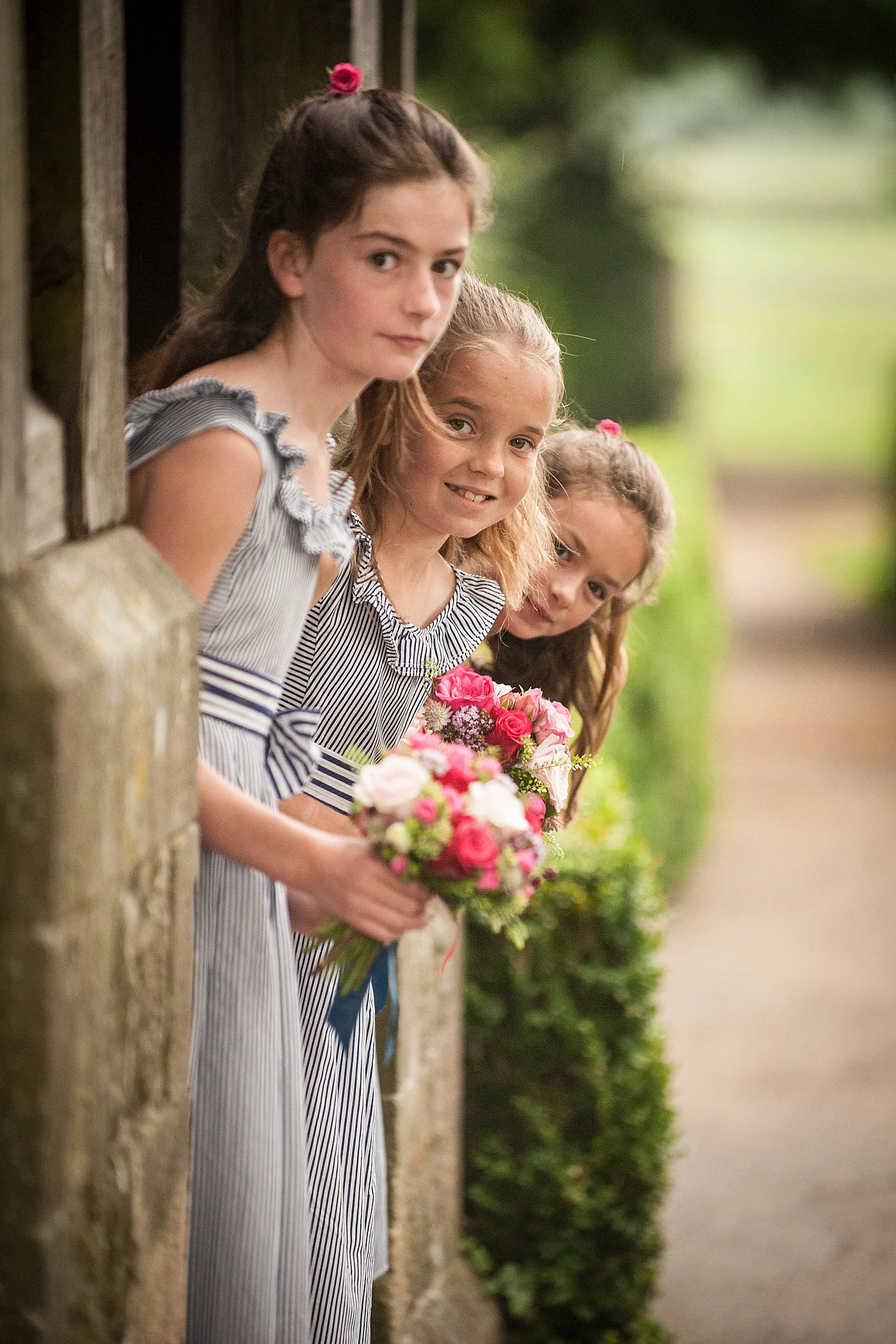 Three young flower girls in striped dresses peek out from a stone doorway holding vibrant pink posy bouquets.