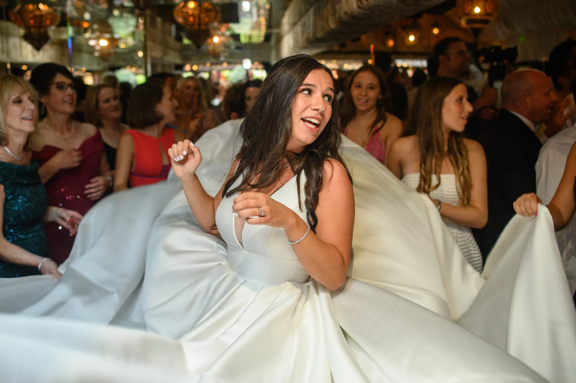 A joyful bride dances energetically on a crowded dance floor, laughing while surrounded by wedding guests holding out her voluminous white satin gown.