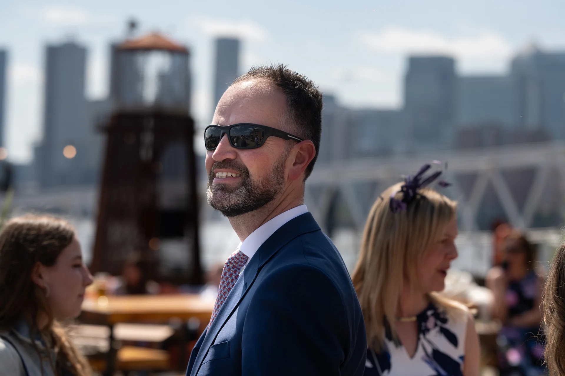 A smiling wedding guest in a navy suit and sunglasses looks away against an urban city skyline and bright sunny sky.