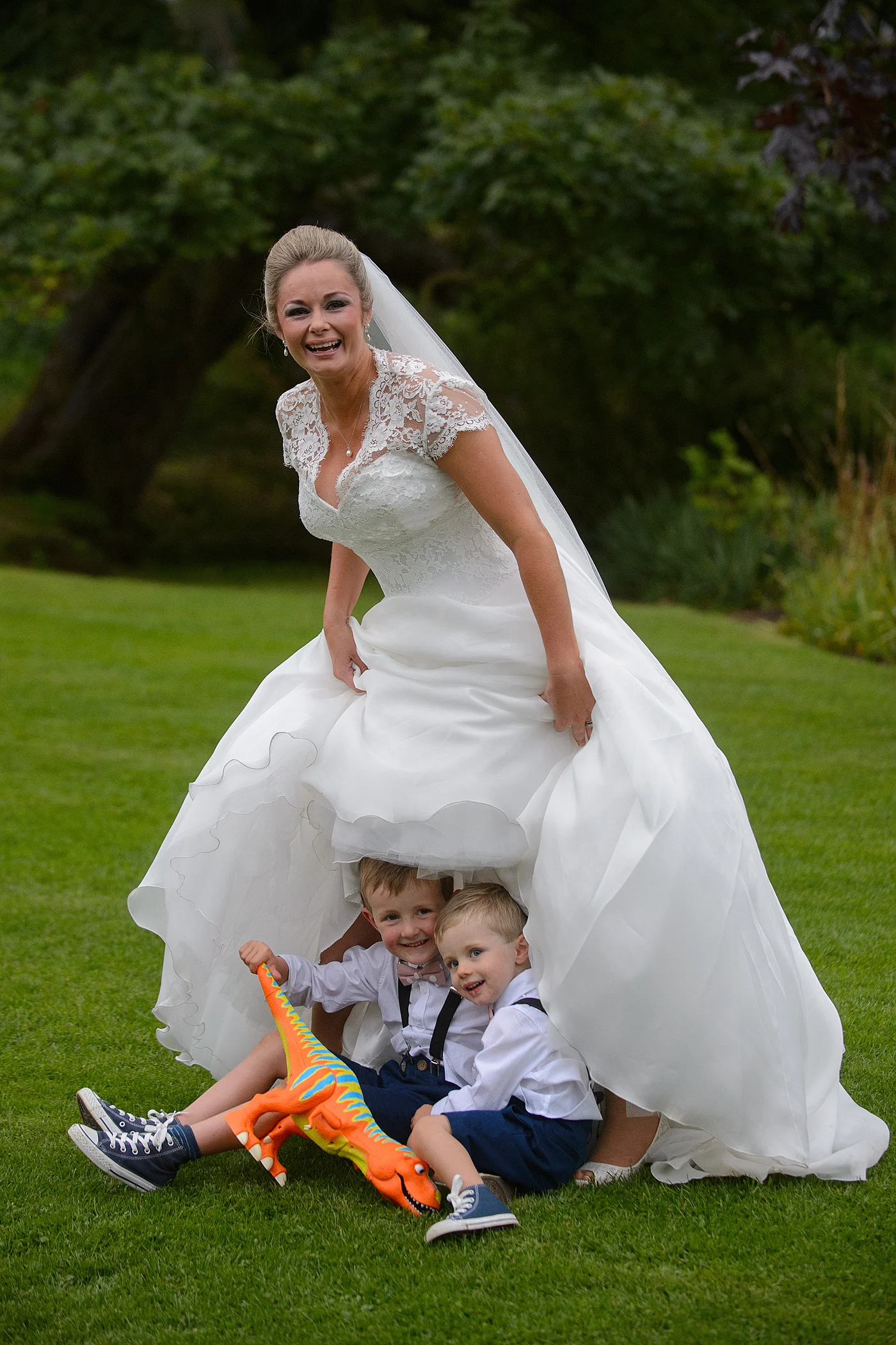 A joyful bride in a lace-detailed a-line wedding gown laughs as she playfully lifts her skirt to reveal two page boys hiding underneath on the grass with a toy dinosaur.