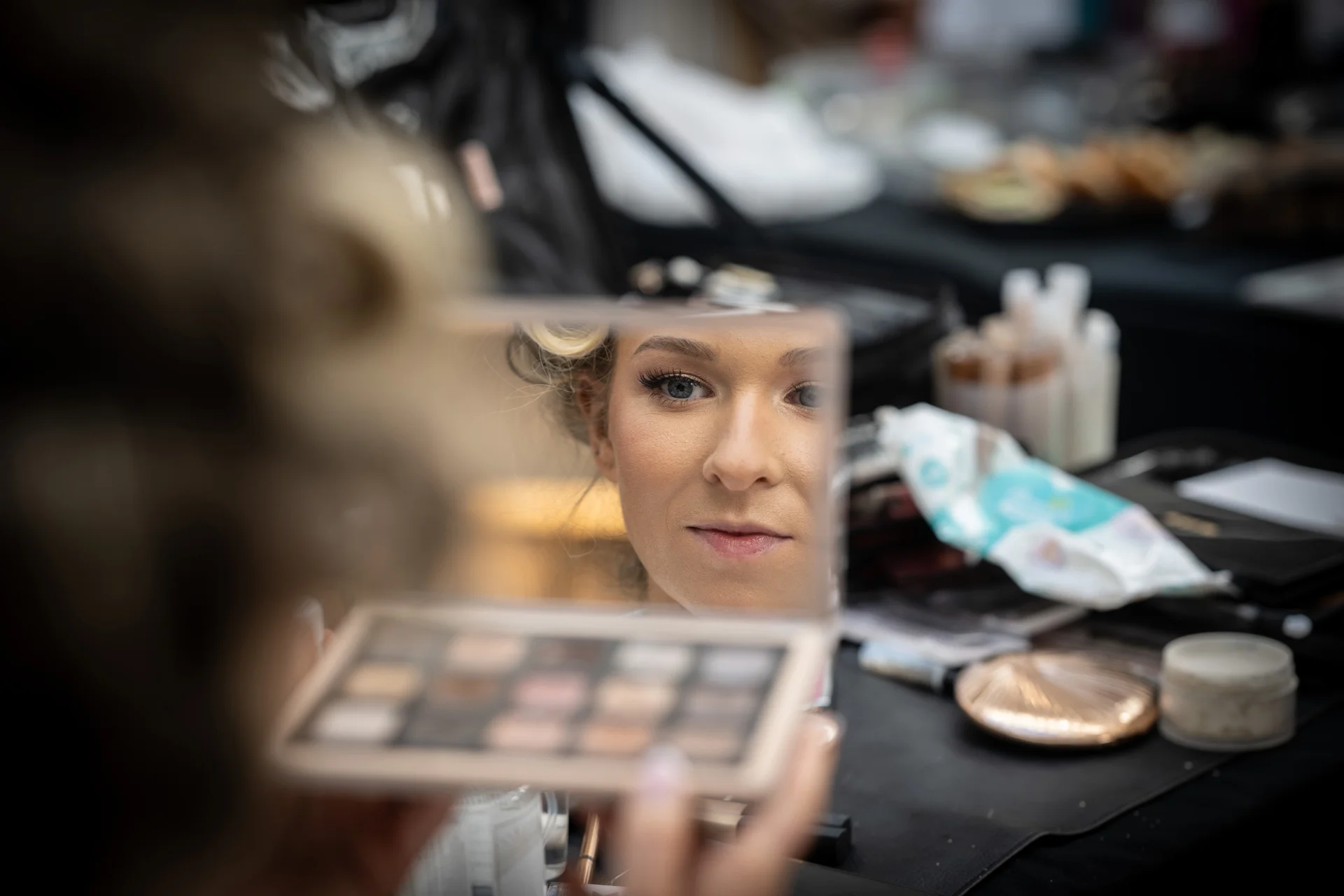 A bride looks into a small makeup mirror while holding a cosmetics palette during her morning bridal preparations.