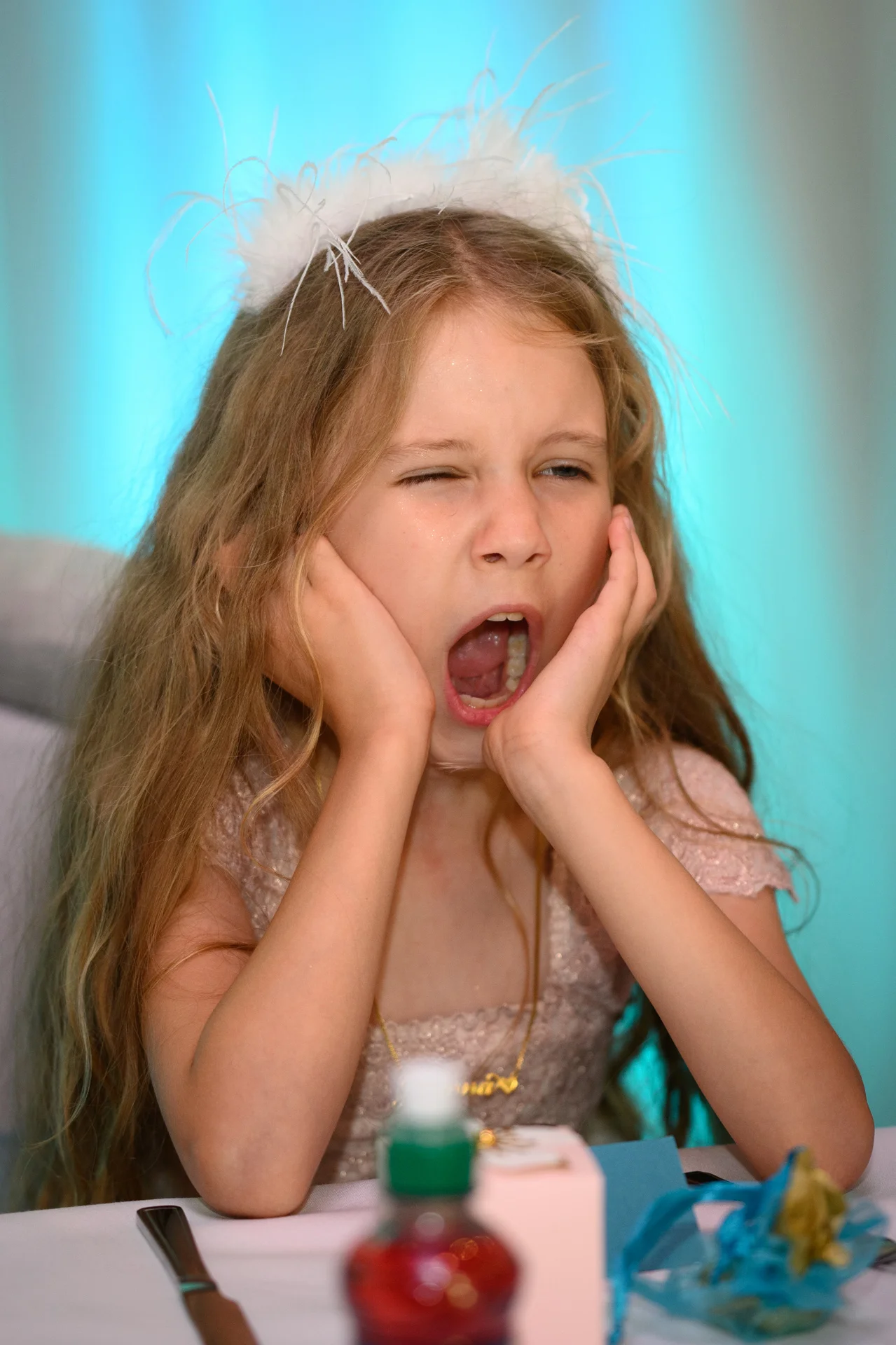 A young flower girl at a wedding reception yawns widely with her hands pressed against her cheeks.