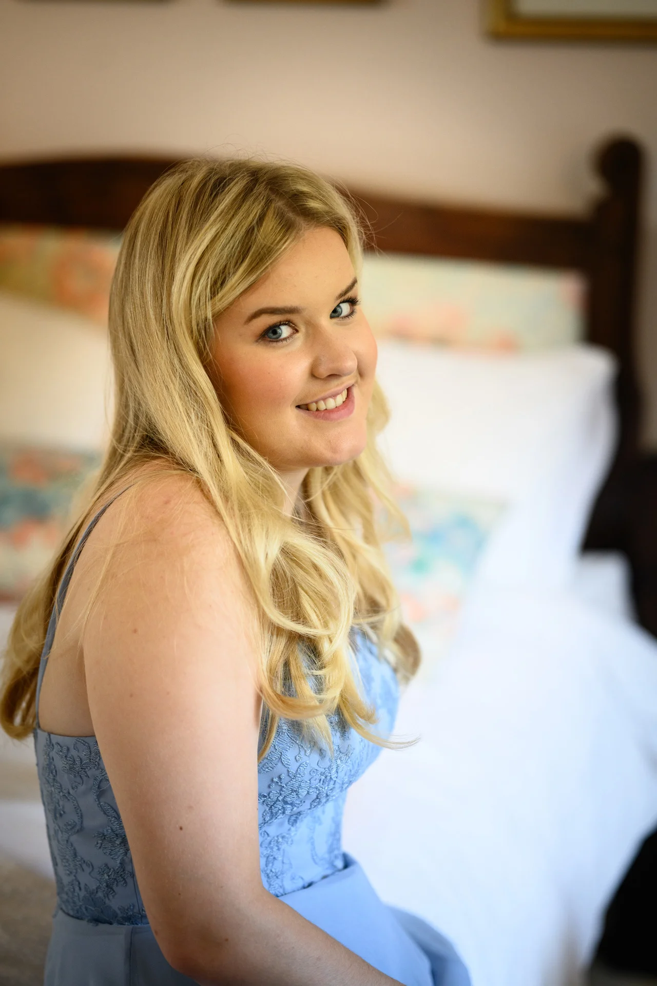 A smiling bridesmaid with blonde hair and a blue dress poses indoors near a bed during wedding morning preparations.