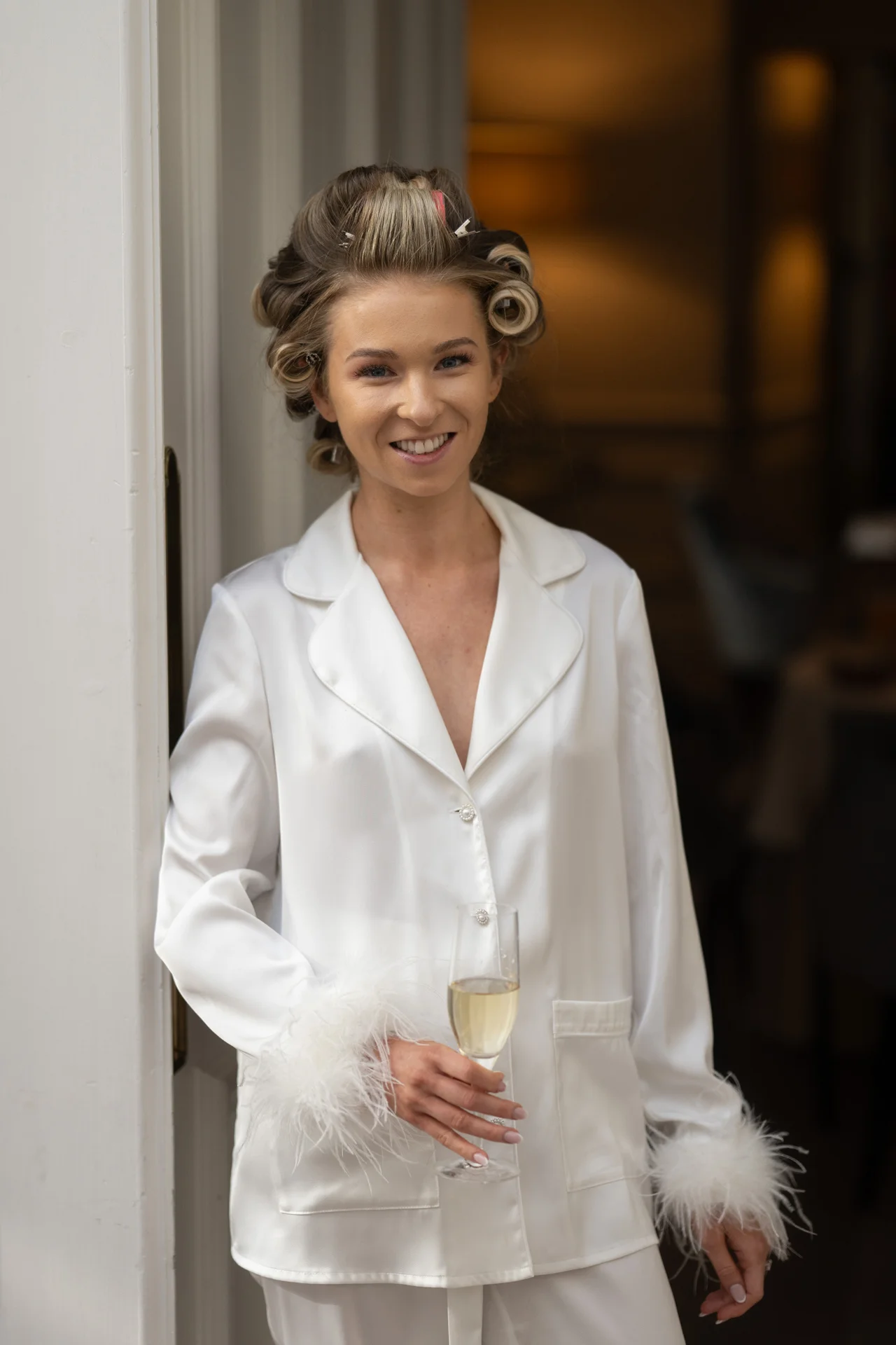 A smiling bride wearing white satin pyjamas with feather-trimmed cuffs poses with a glass of champagne during morning wedding preparations.