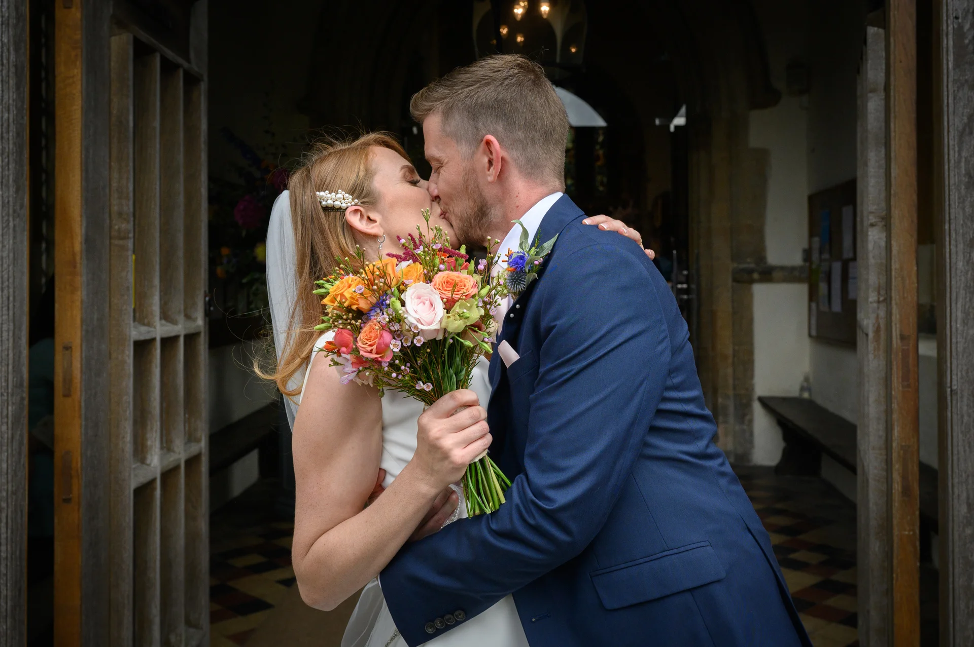 A newlywed couple share their first kiss outside a church doorway, with the bride holding a vibrant, colourful bouquet of flowers.