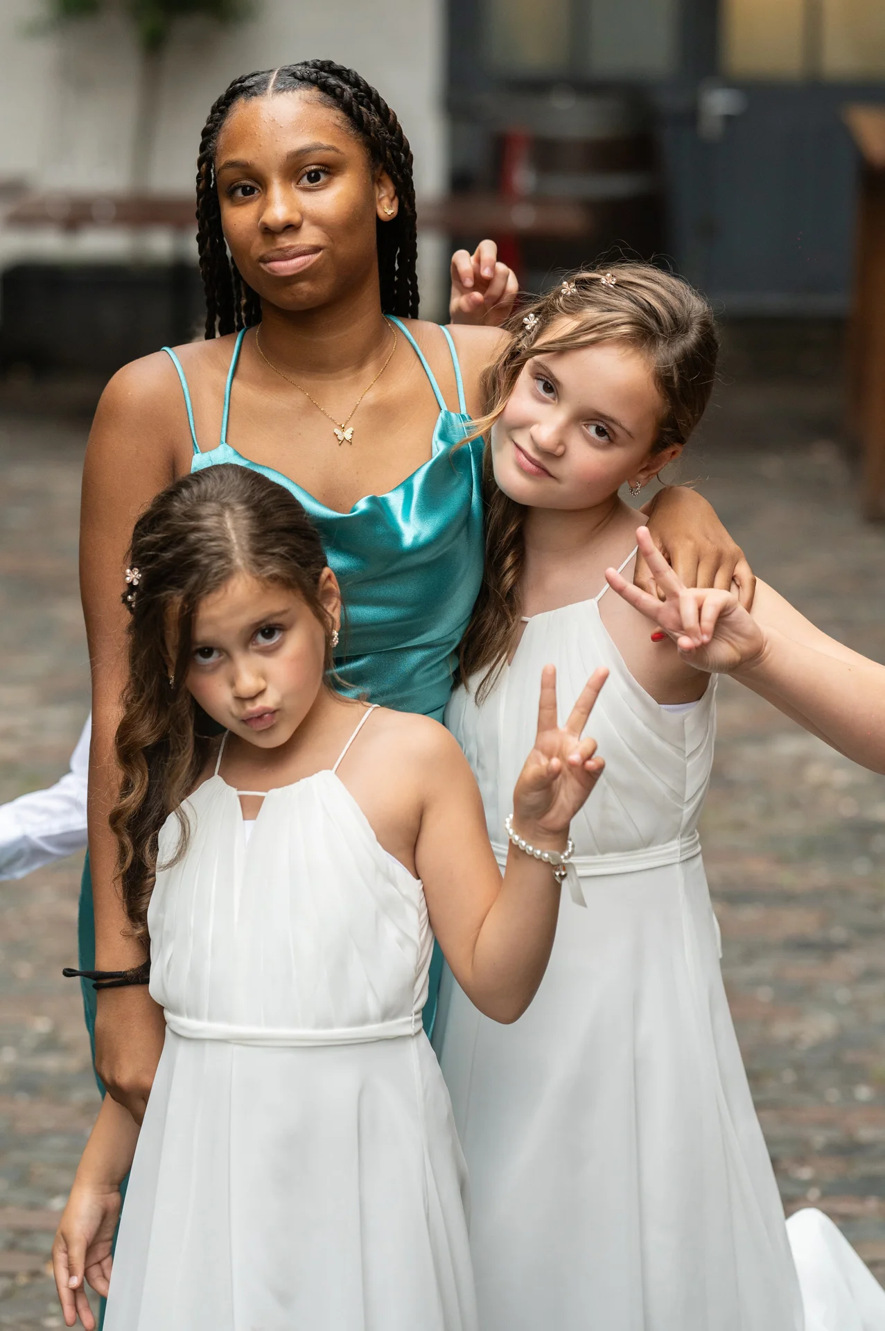 Three young wedding attendants pose playfully together in a cobbled courtyard, wearing white dresses and a turquoise slip dress while flashing peace signs.