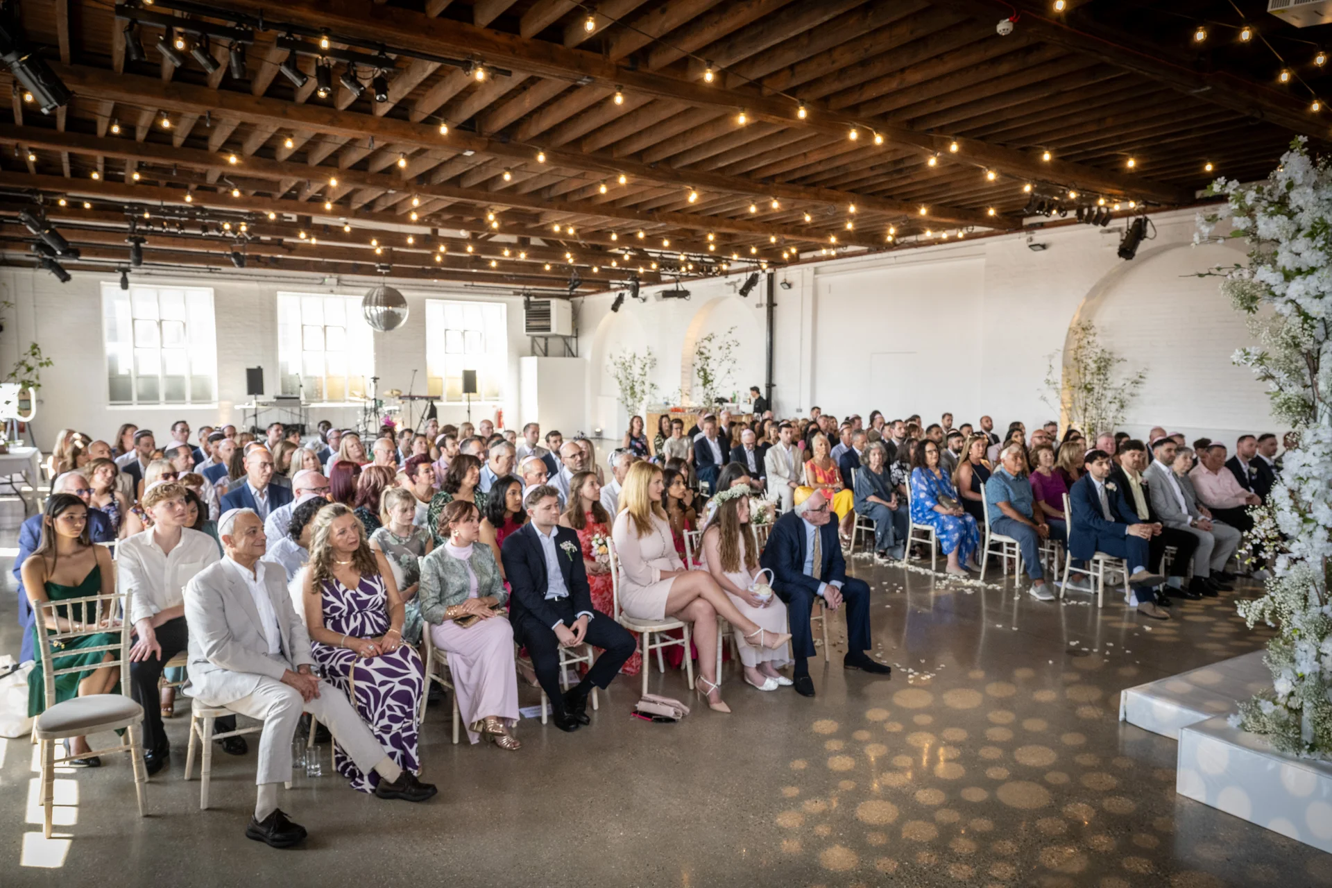 A wide angle shot of a warehouse wedding ceremony featuring guests seated under exposed wooden rafters and a glowing fairy light canopy.