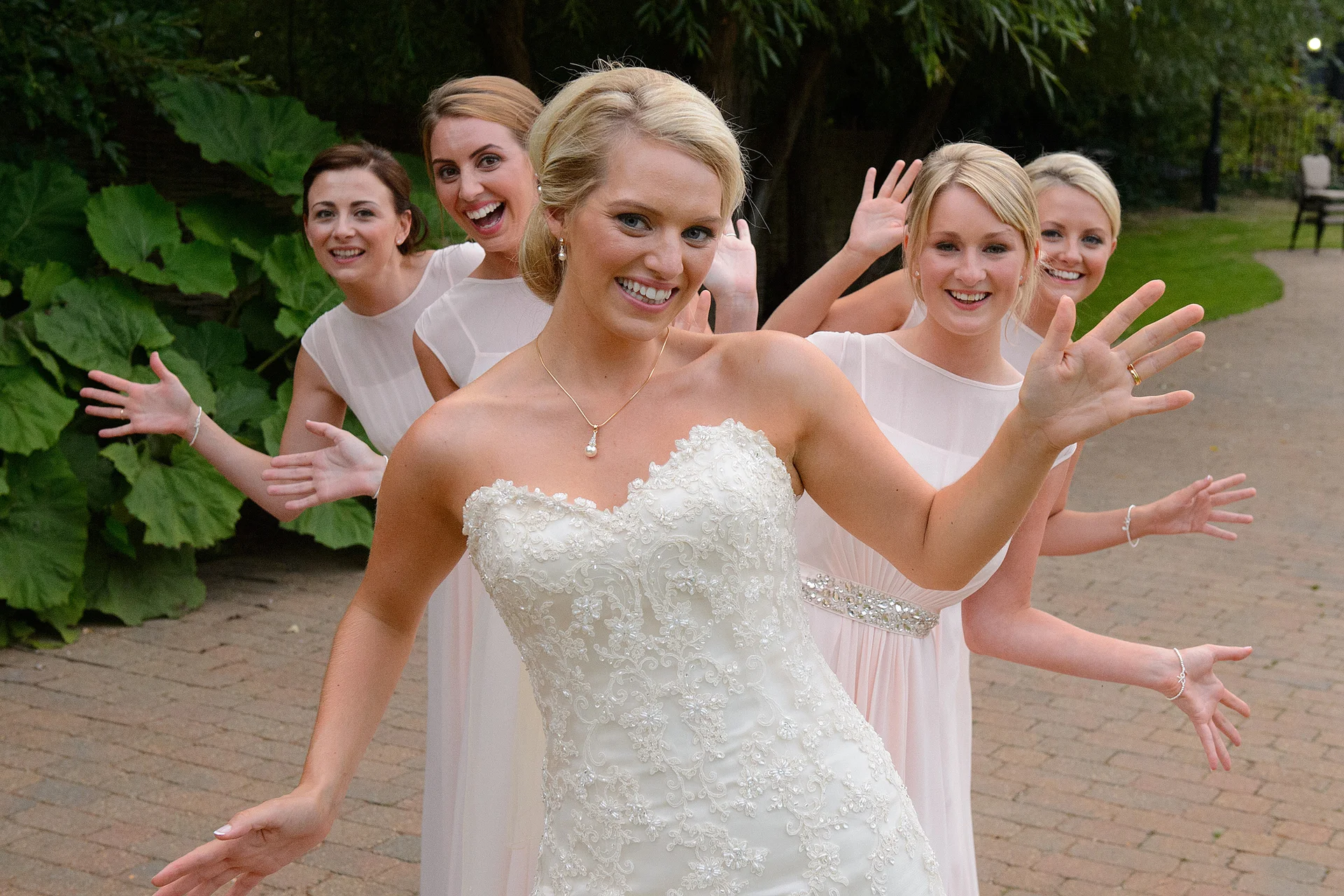 A smiling bride in a beaded strapless wedding dress poses playfully with her four bridesmaids wearing matching pink gowns outdoors.