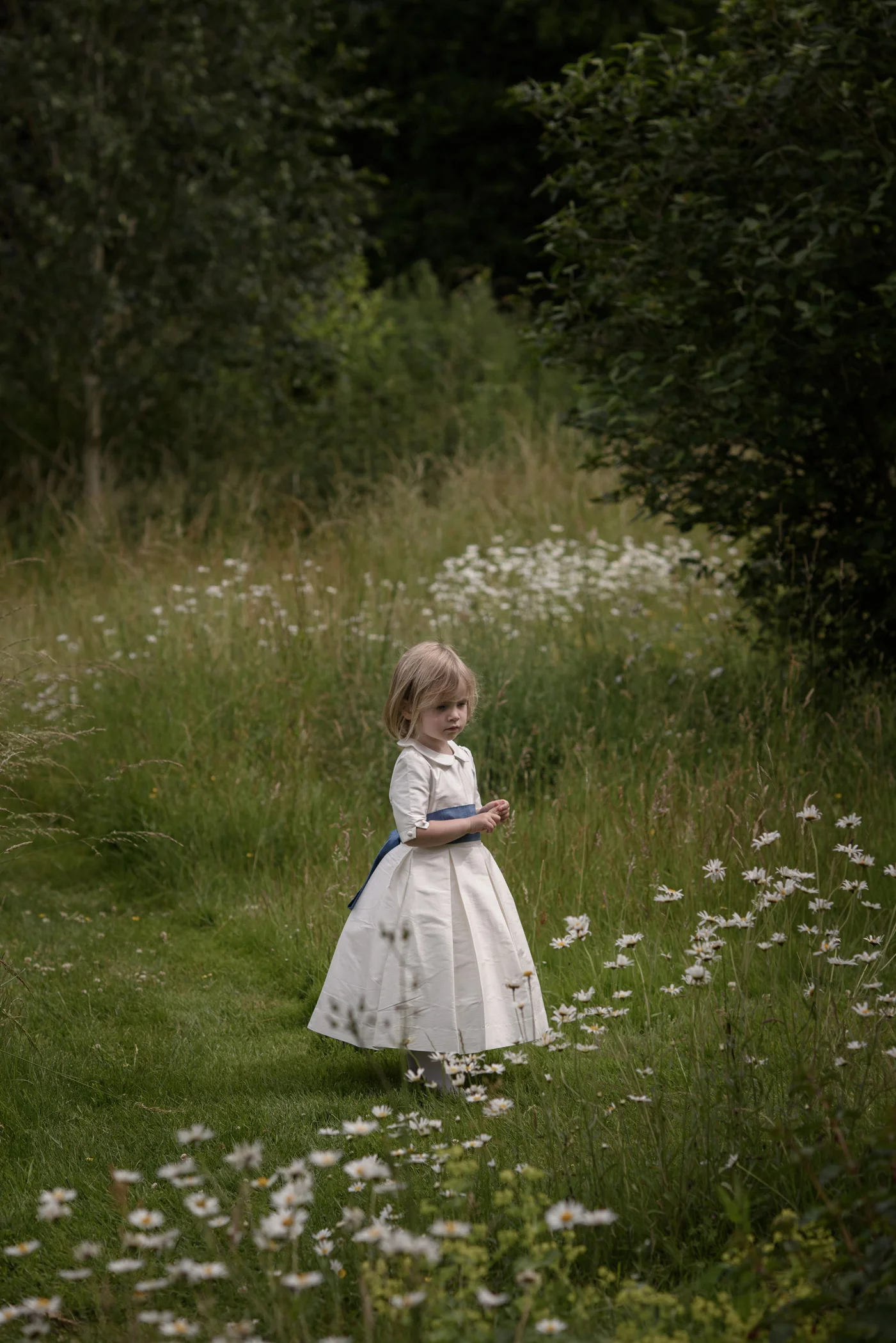 A young flower girl in a white dress stands quietly amongst a lush wildflower meadow during an outdoor woodland wedding.
