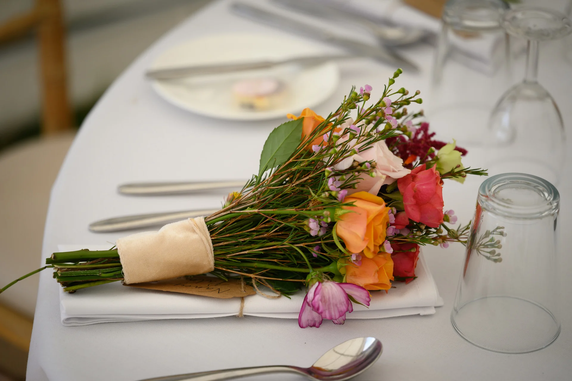 A delicate hand-tied bouquet featuring orange and pink roses rests neatly on a crisp white napkin atop a wedding reception table.