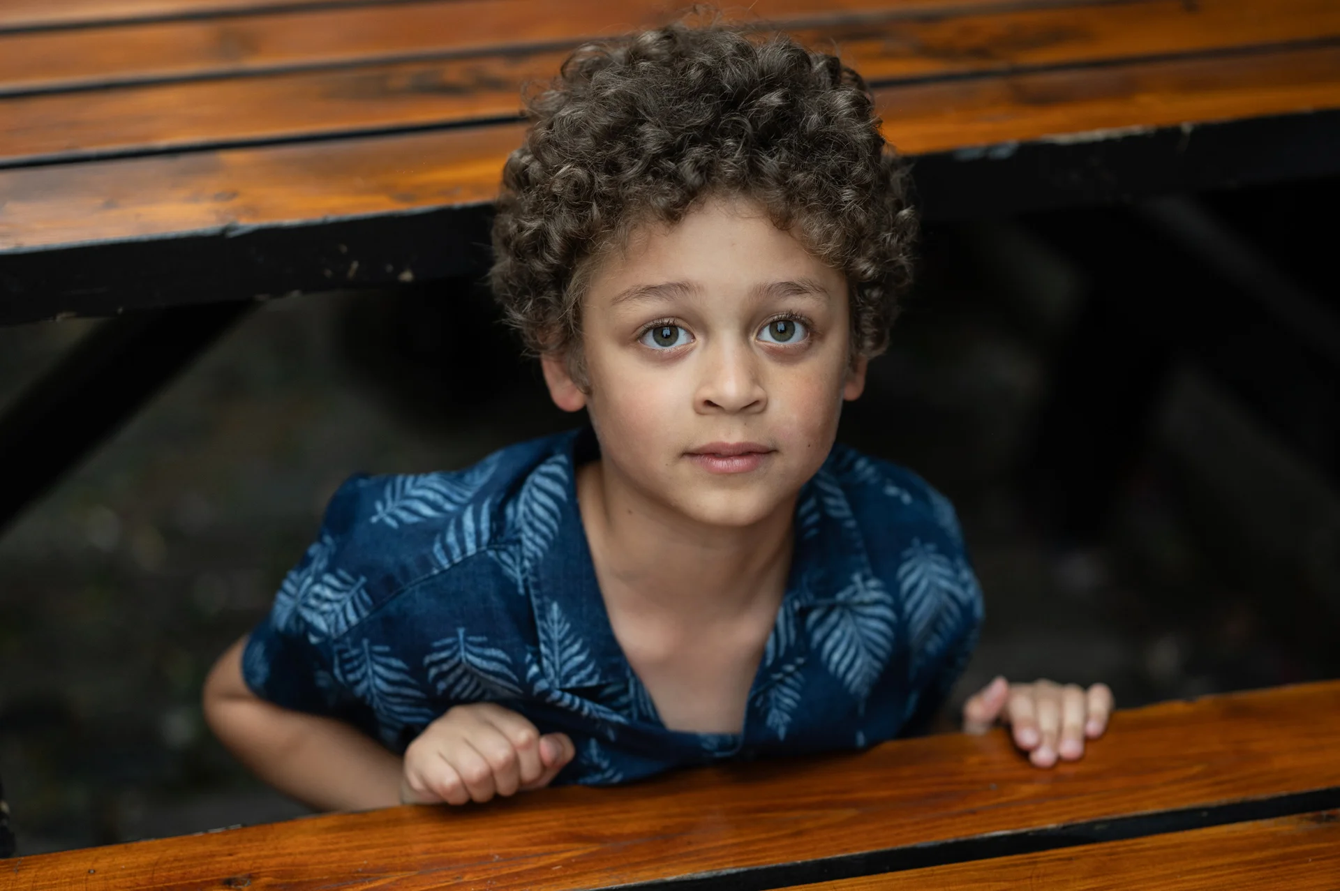 A young child with curly hair looks up curiously from beneath a polished wooden table at a wedding celebration.