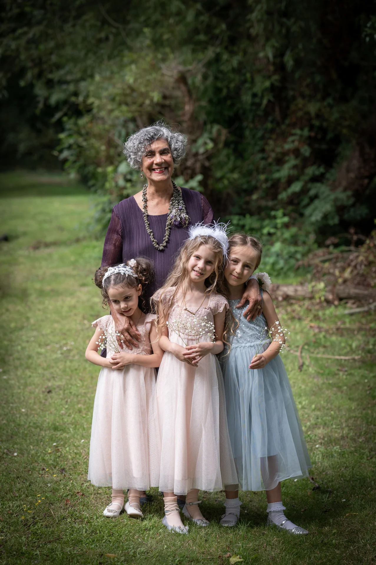 An older woman with curly grey hair smiles while posing outdoors in a woodland setting with three young flower girls in tulle dresses holding tiny stems of gypsophila.