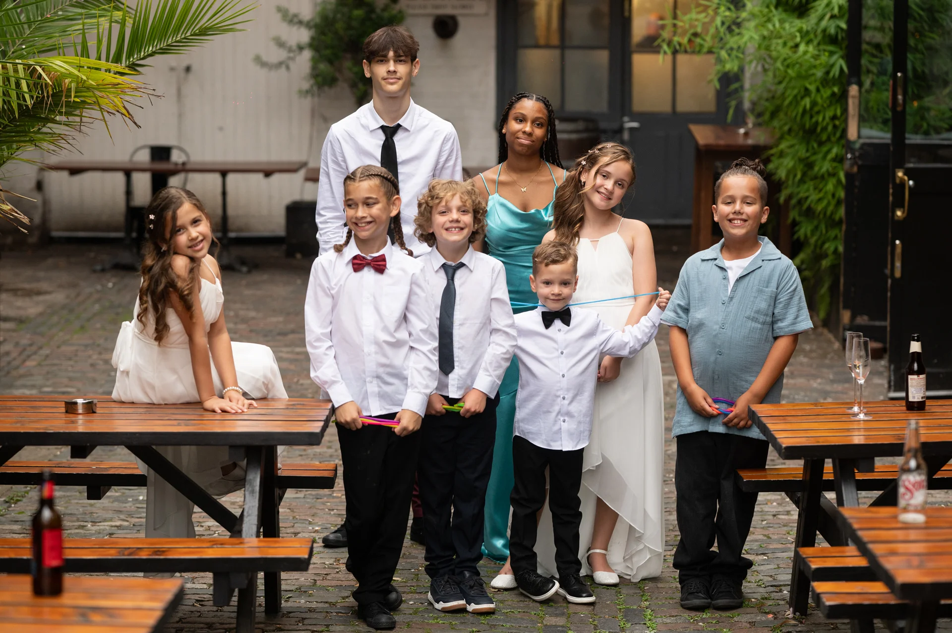 A group of young wedding guests and children pose together in an outdoor courtyard setting near wooden picnic tables.