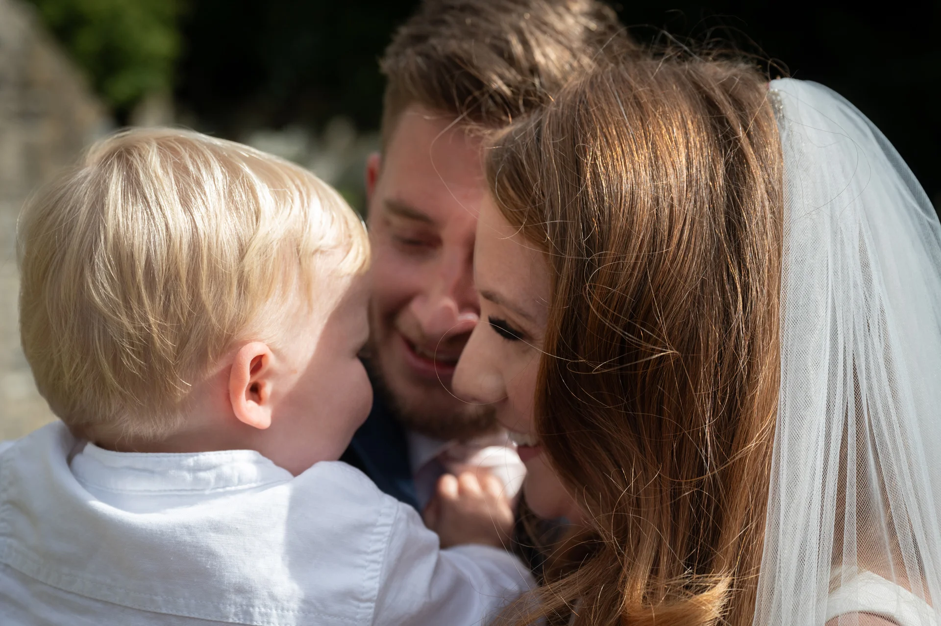 A bride and groom share a tender moment outdoors while holding a young child wearing a white shirt.