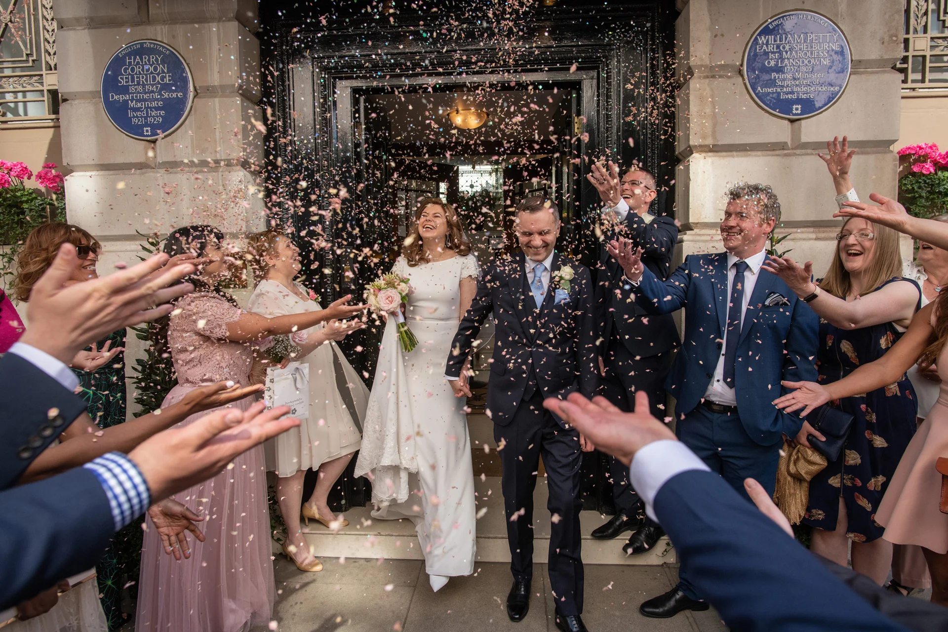 A joyful newlywed couple laughs and walks hand in hand out of a historic London venue as family and guests enthusiastically throw colourful confetti over them.