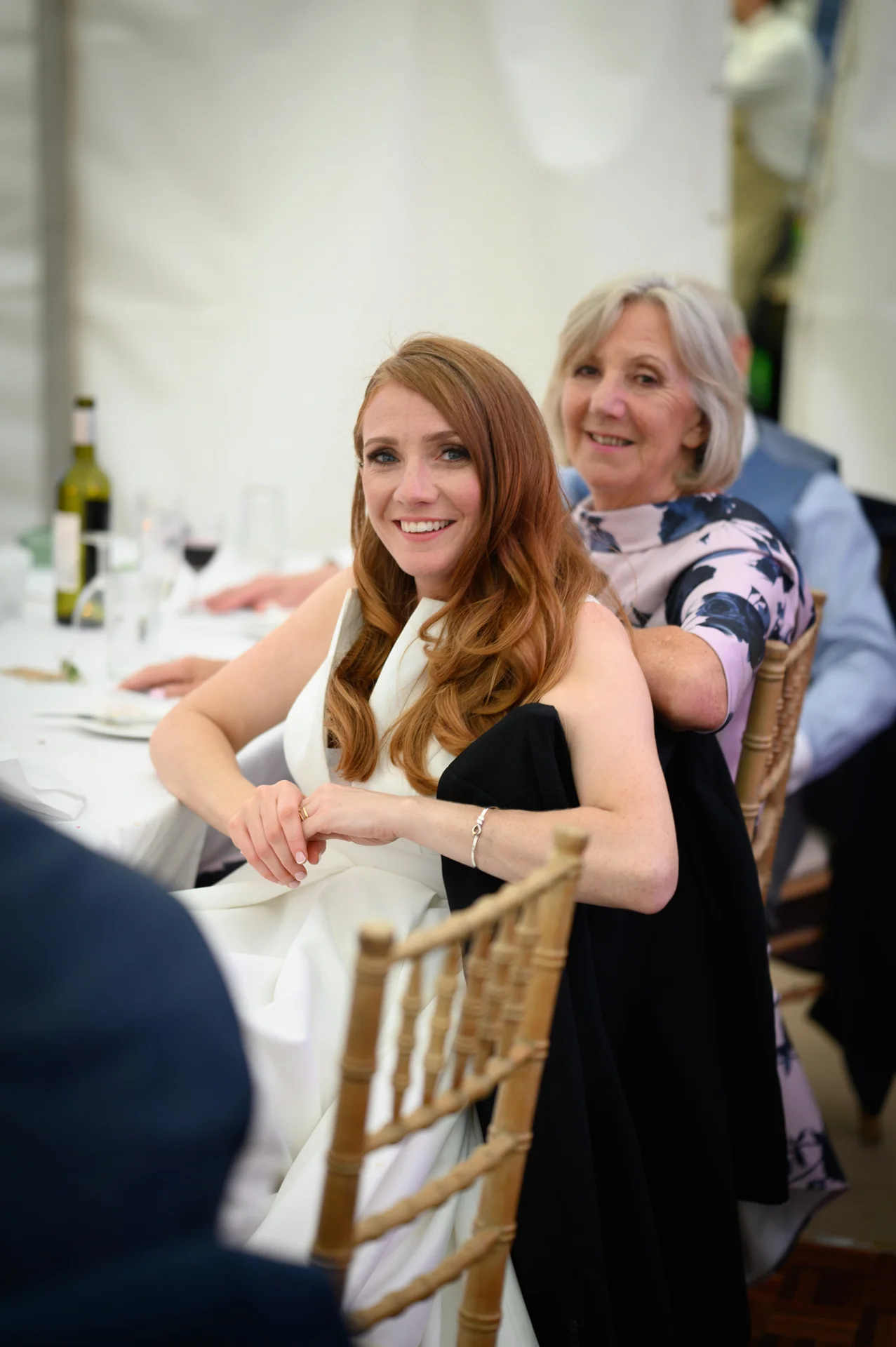 A smiling bride with long red hair sits at a wedding breakfast table inside a marquee, accompanied by guests.