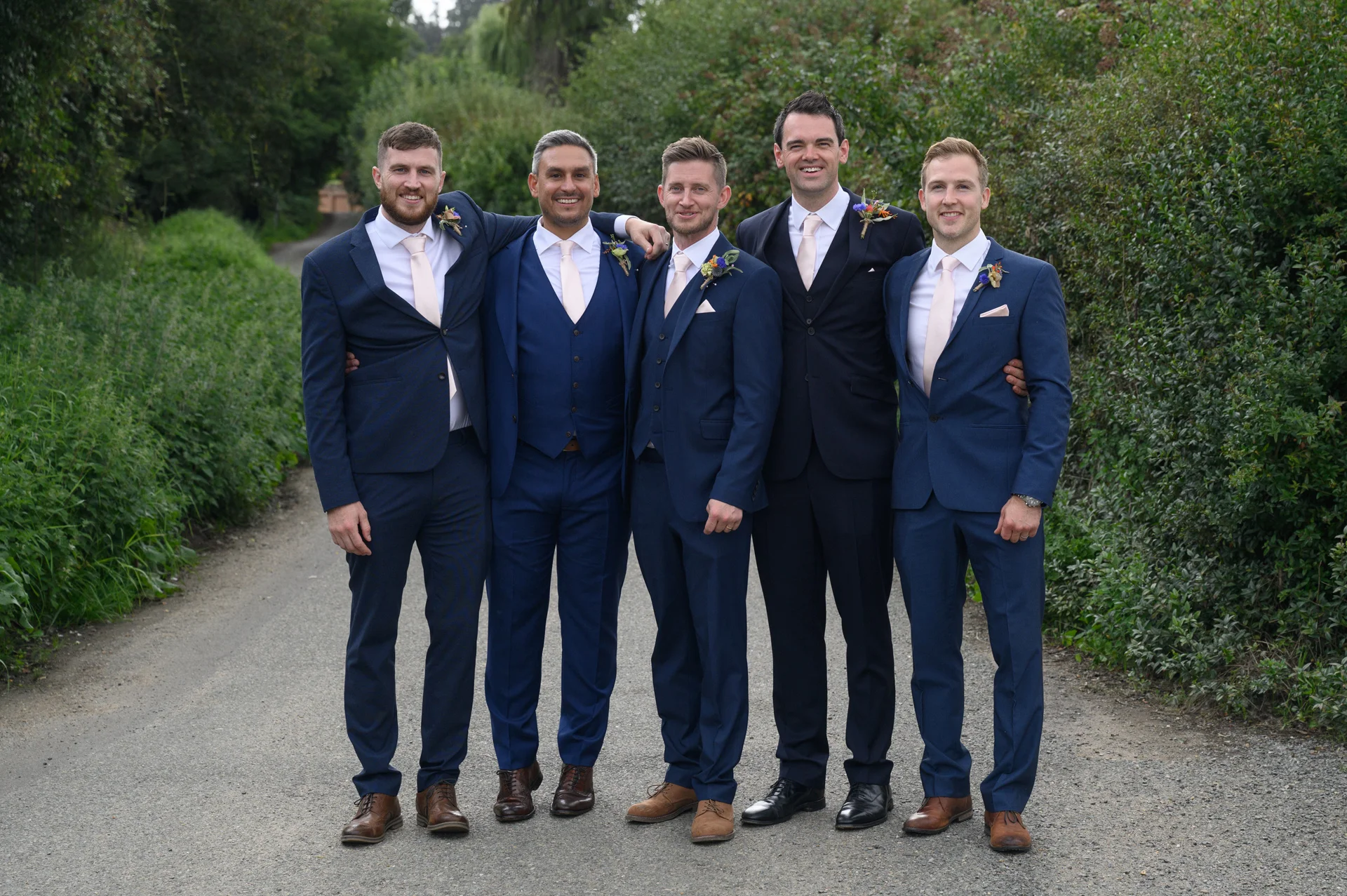 A group of five smiling groomsmen in matching navy suits and pink ties pose together along a leafy country lane.