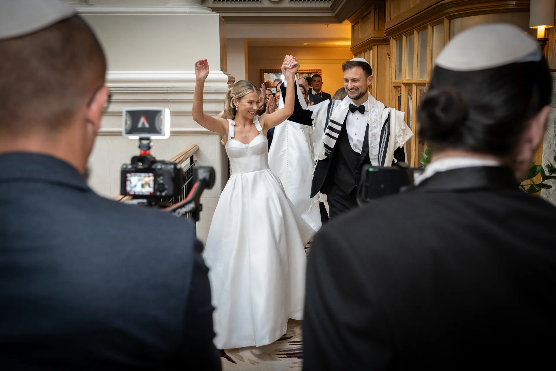 A newlywed couple joyfully walks hand in hand with arms raised amidst cheering guests wearing kippahs, captured in a documentary style.