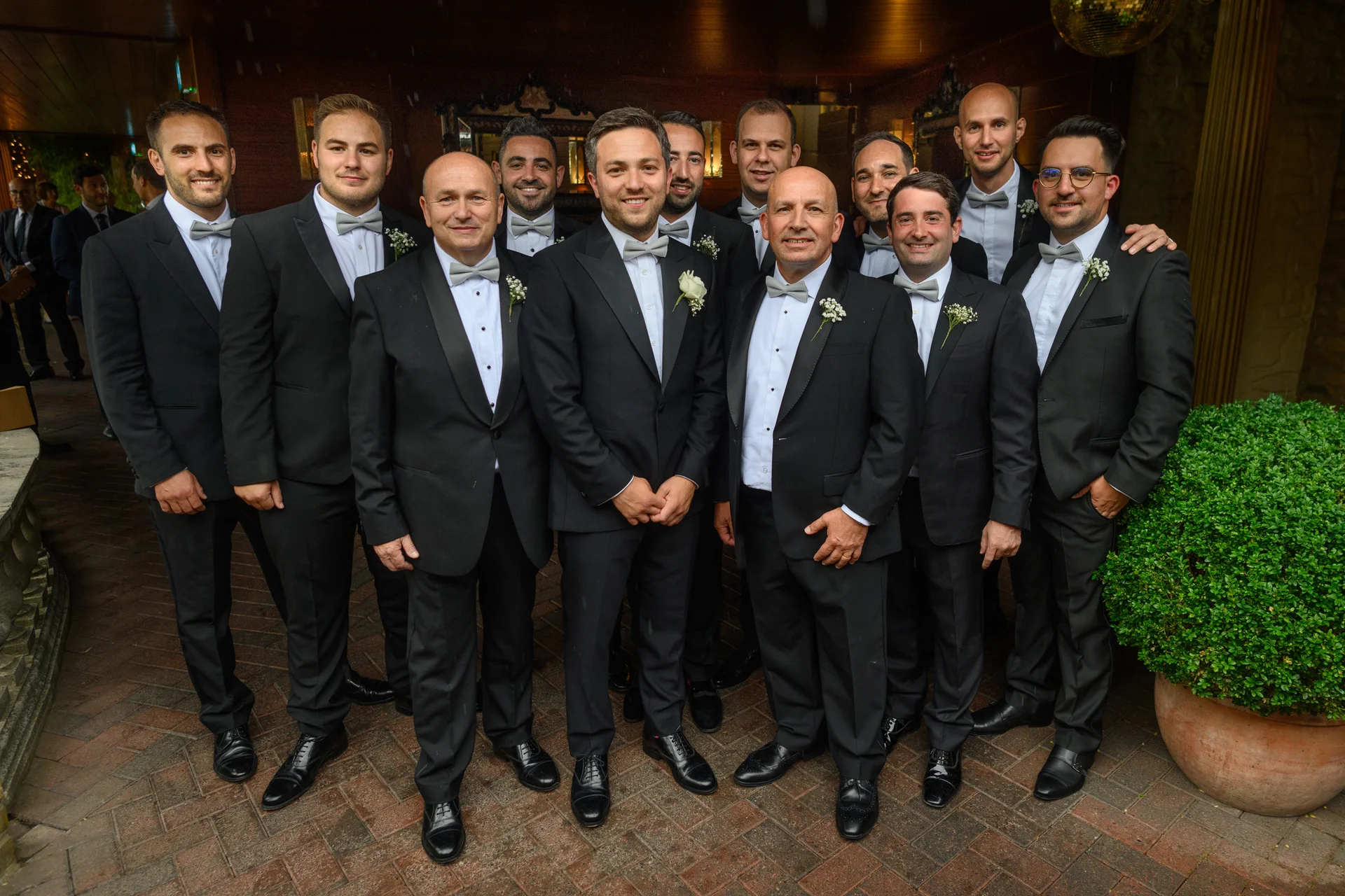 A large group of groomsmen dressed in classic black tuxedos with bow ties and white buttonholes pose together outdoors on a brick patio.