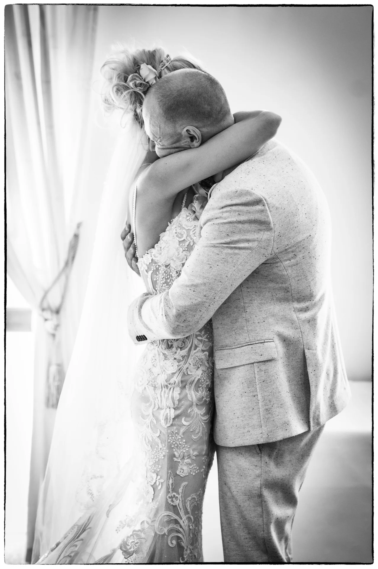 A newlywed couple shares an emotional embrace in soft window light, with the bride wearing an intricate lace gown and the groom in a textured suit jacket.