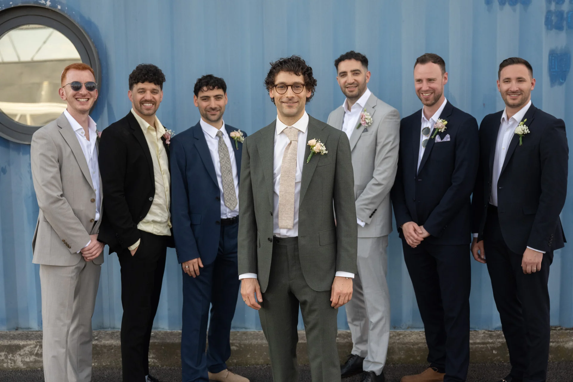 A smiling groom in an olive green suit with a textured tie stands centrally in front of a blue industrial wall, flanked by five groomsmen wearing mismatched suits with floral buttonholes.
