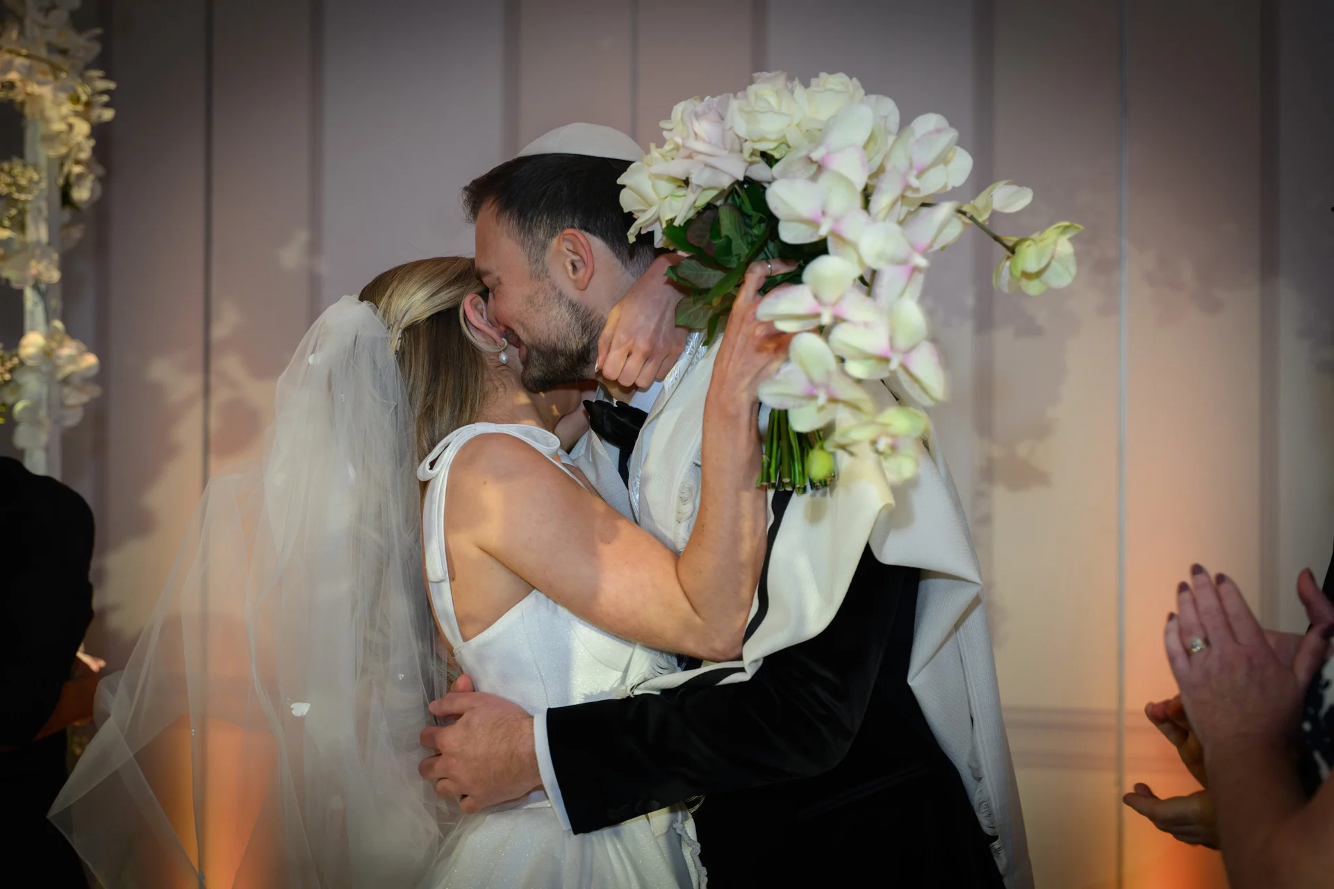 The bride and groom share an emotional first kiss under a traditional Jewish wedding canopy while holding an oversized orchid bouquet.