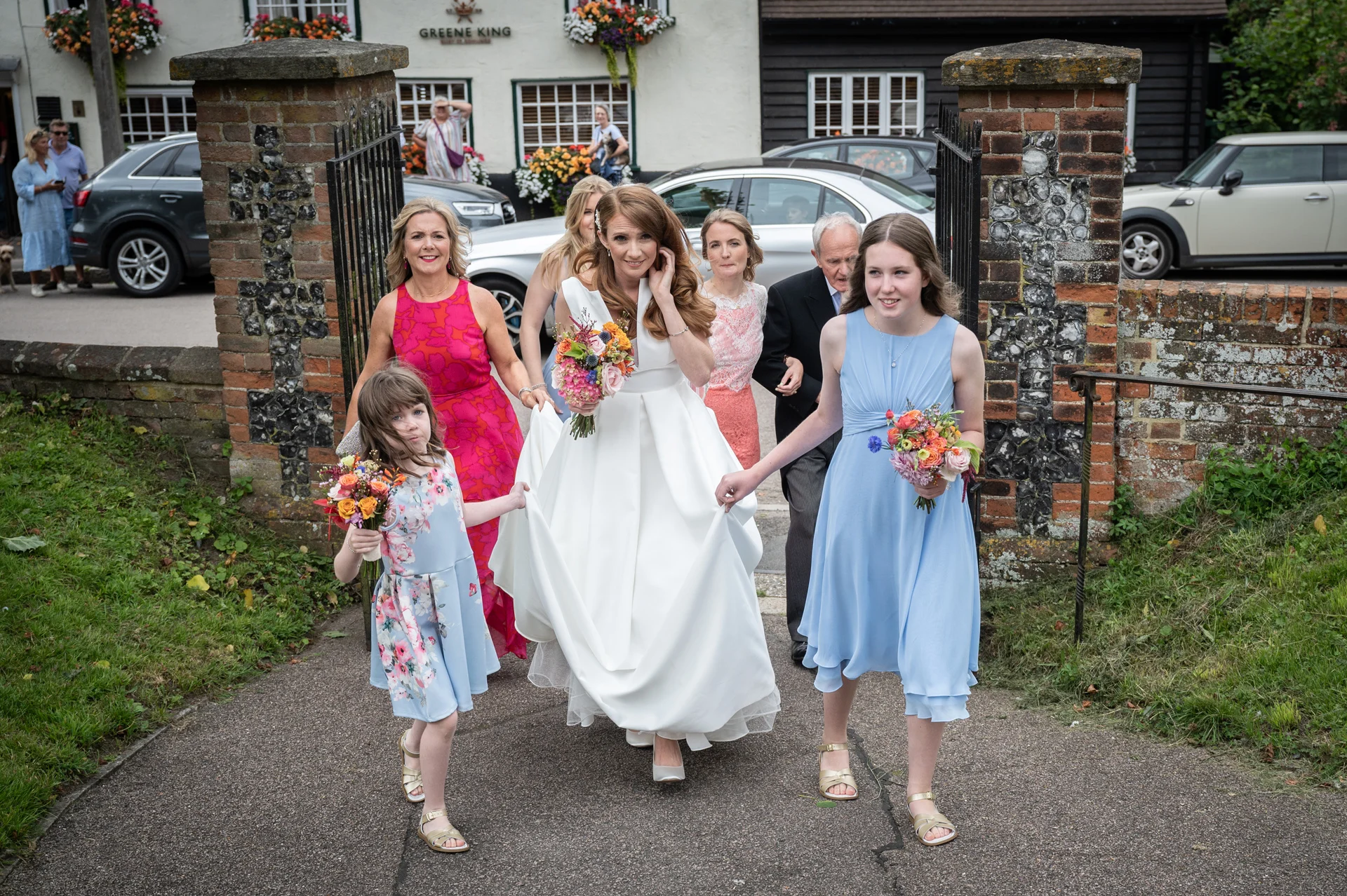 A smiling bride holding a colourful bouquet walks through brick gate pillars accompanied by her bridesmaids, flower girl, and father.