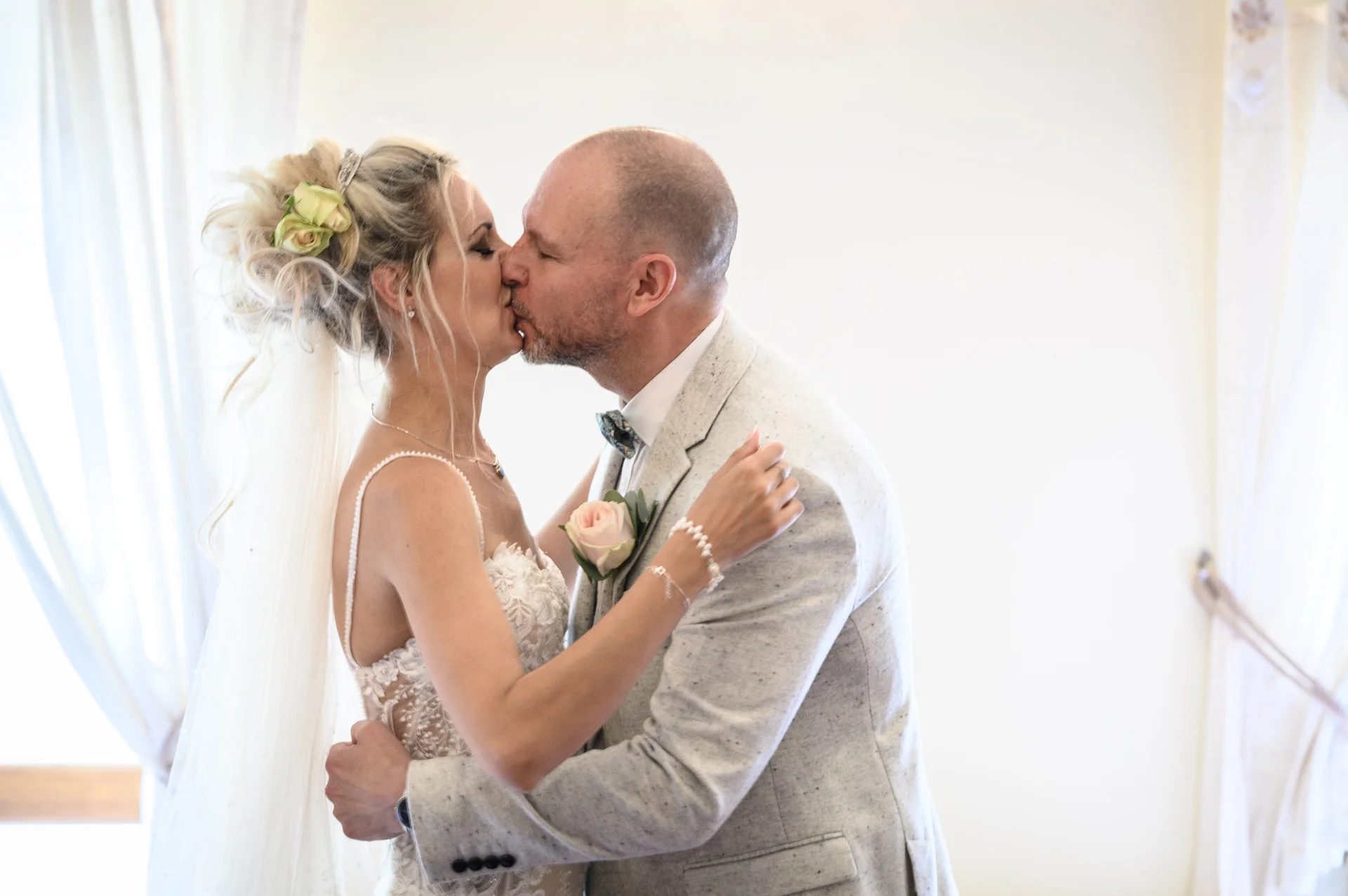 The newly married couple share a tender kiss indoors near a brightly lit window, with the bride wearing a textured updo and a delicate spaghetti strap gown.
