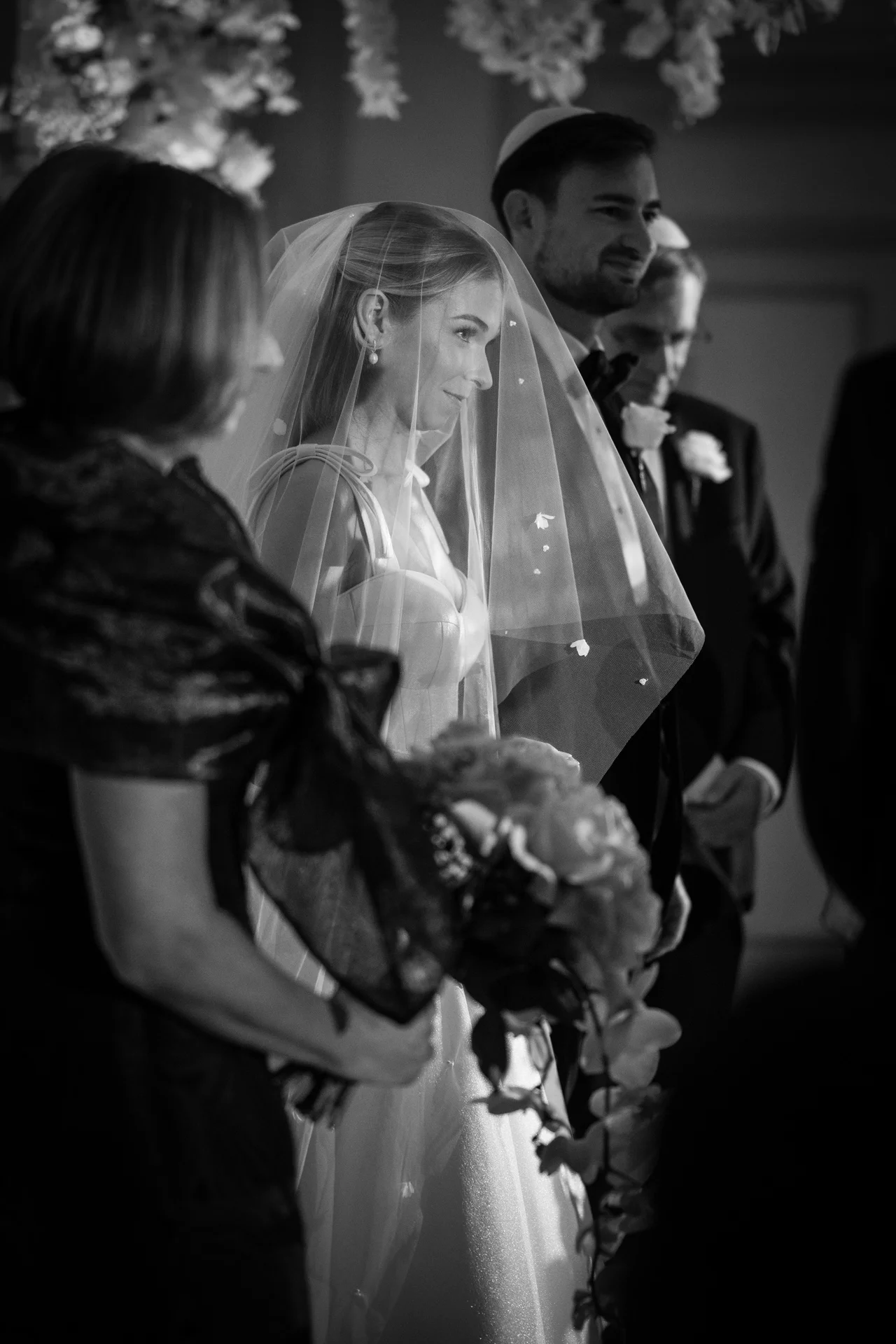 An emotional black and white shot of a bride wearing a blusher veil and holding a floral bouquet, standing at the altar beside the groom and family members during a traditional ceremony.