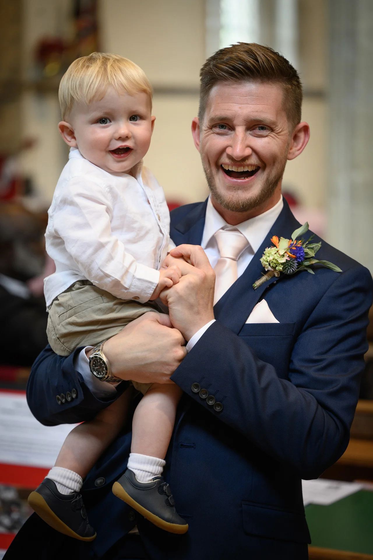 A smiling groom wearing a navy suit and floral buttonhole happily holds a young blond child in his arms.