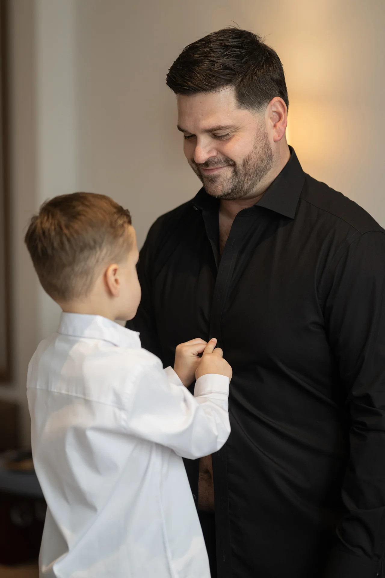 A young page boy in a crisp white shirt smiling as he helps an adult groom in a black shirt get dressed on the morning of the wedding.