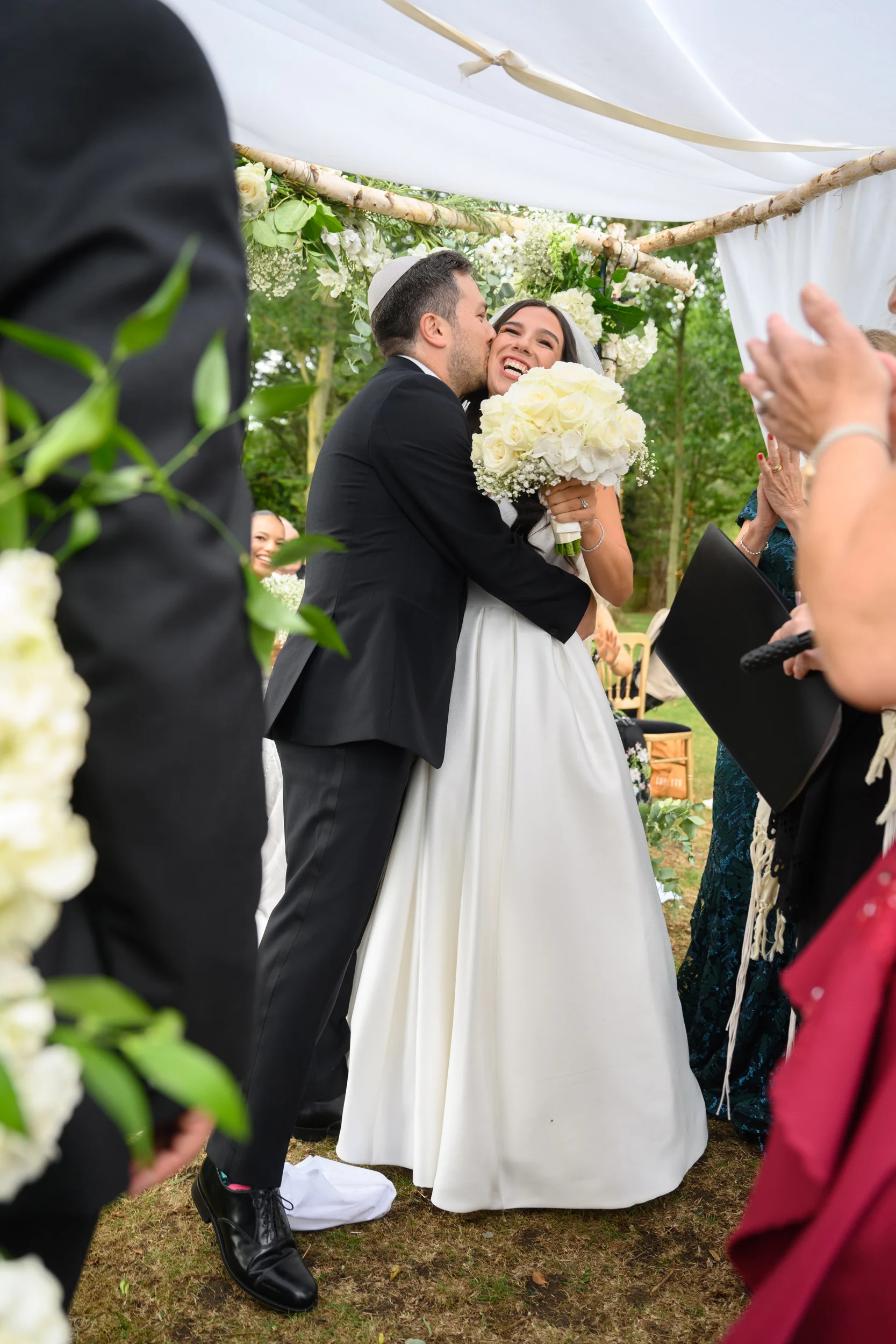 A newly married couple share an emotional embrace and kiss beneath an outdoor chuppah adorned with white roses during their wedding ceremony.