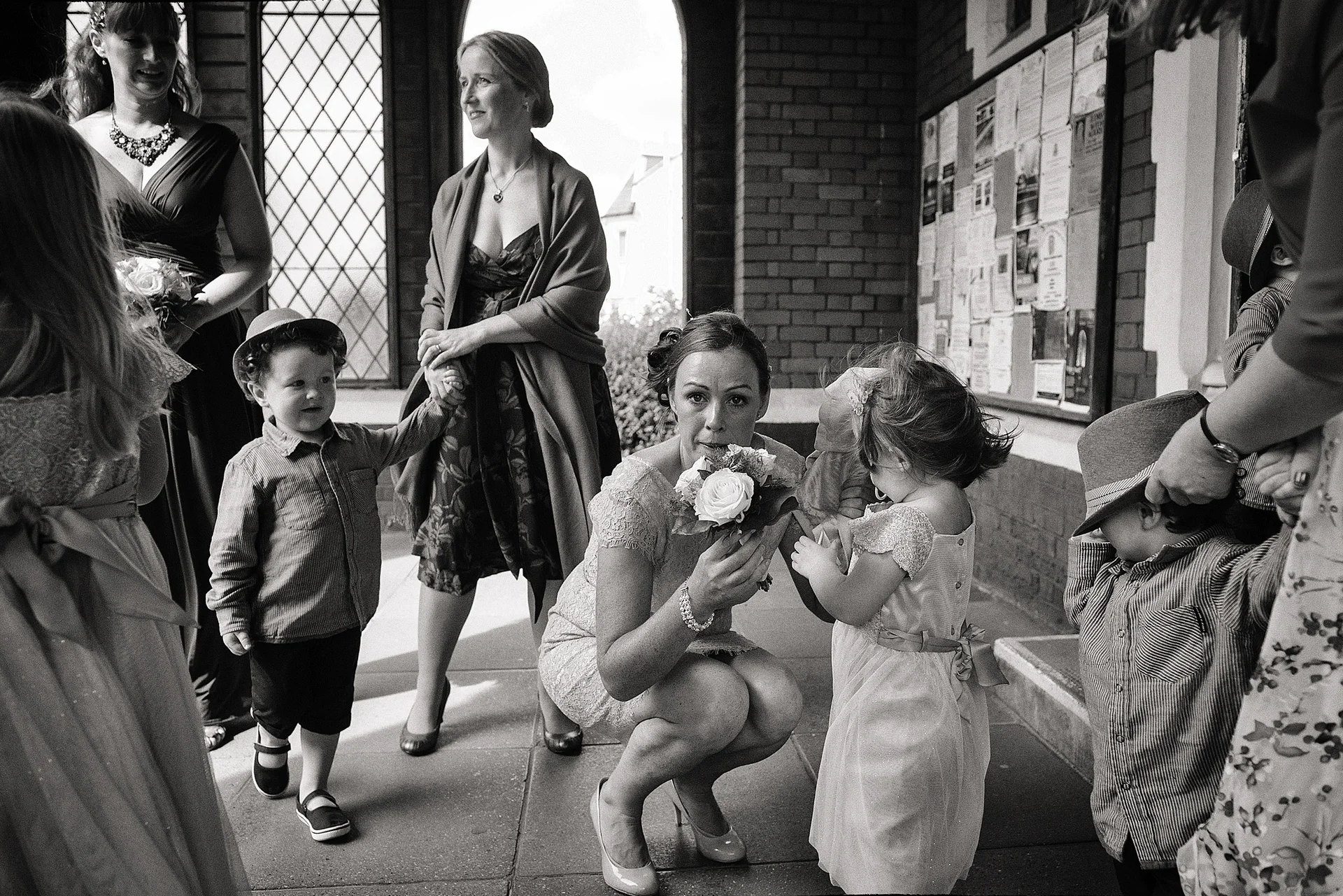 A candid black and white shot captures guests and children gathered near a brick doorway, with a woman crouching down to interact with a young flower girl holding a small bouquet.