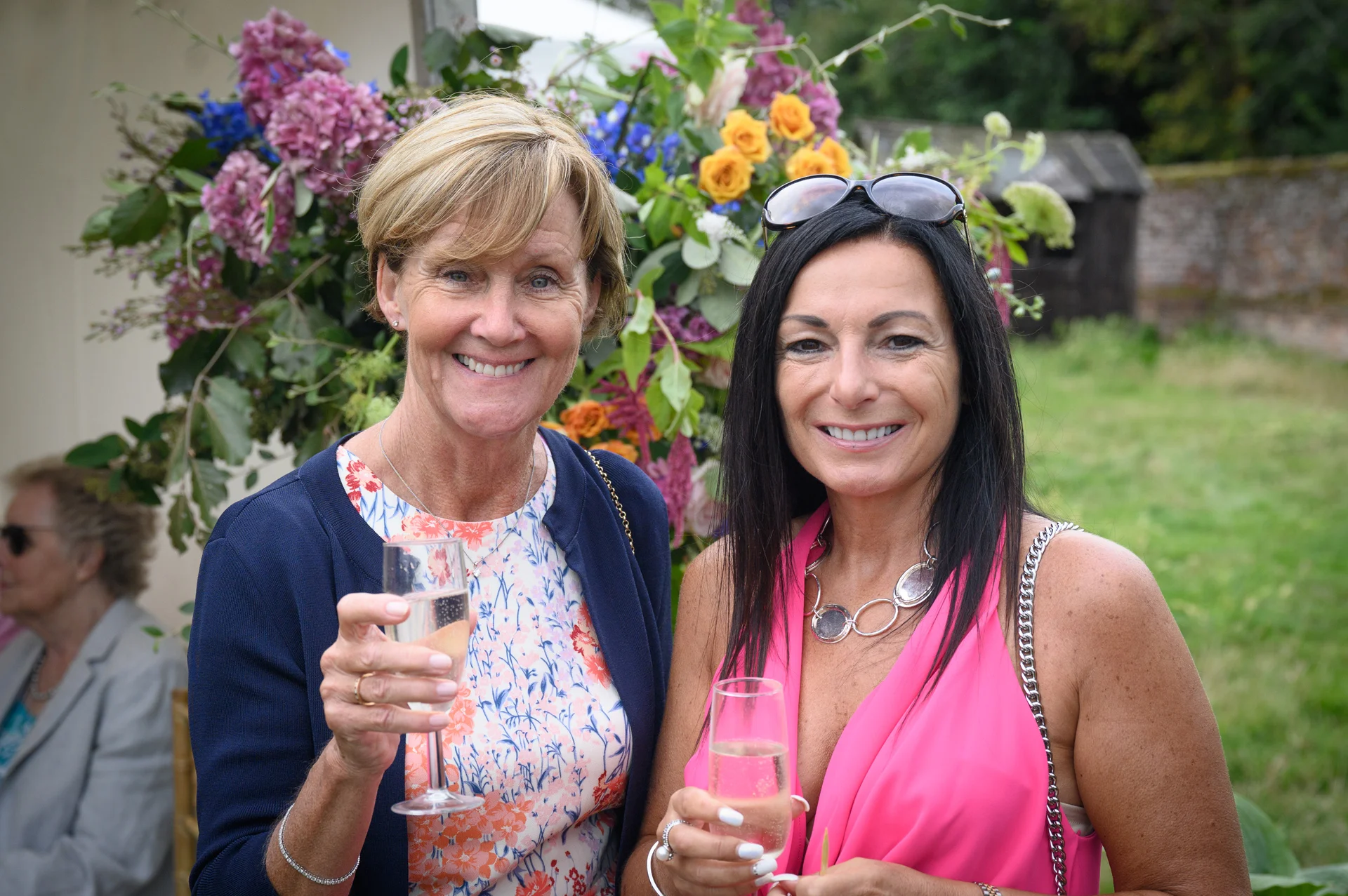 Two smiling female wedding guests hold champagne flutes and pose outdoors in front of a colourful floral arch during the drinks reception.
