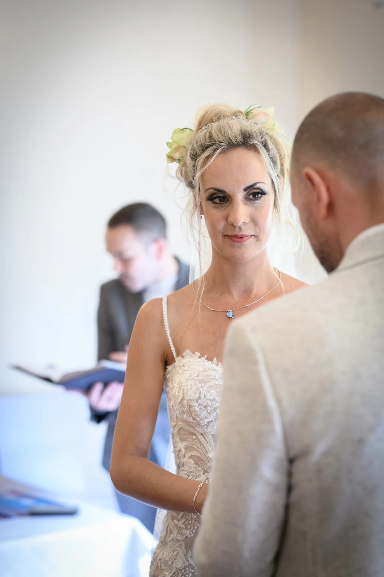 A bride in a lace gown looks lovingly at her groom during their intimate civil ceremony, with the registrar visible in the softly blurred background.