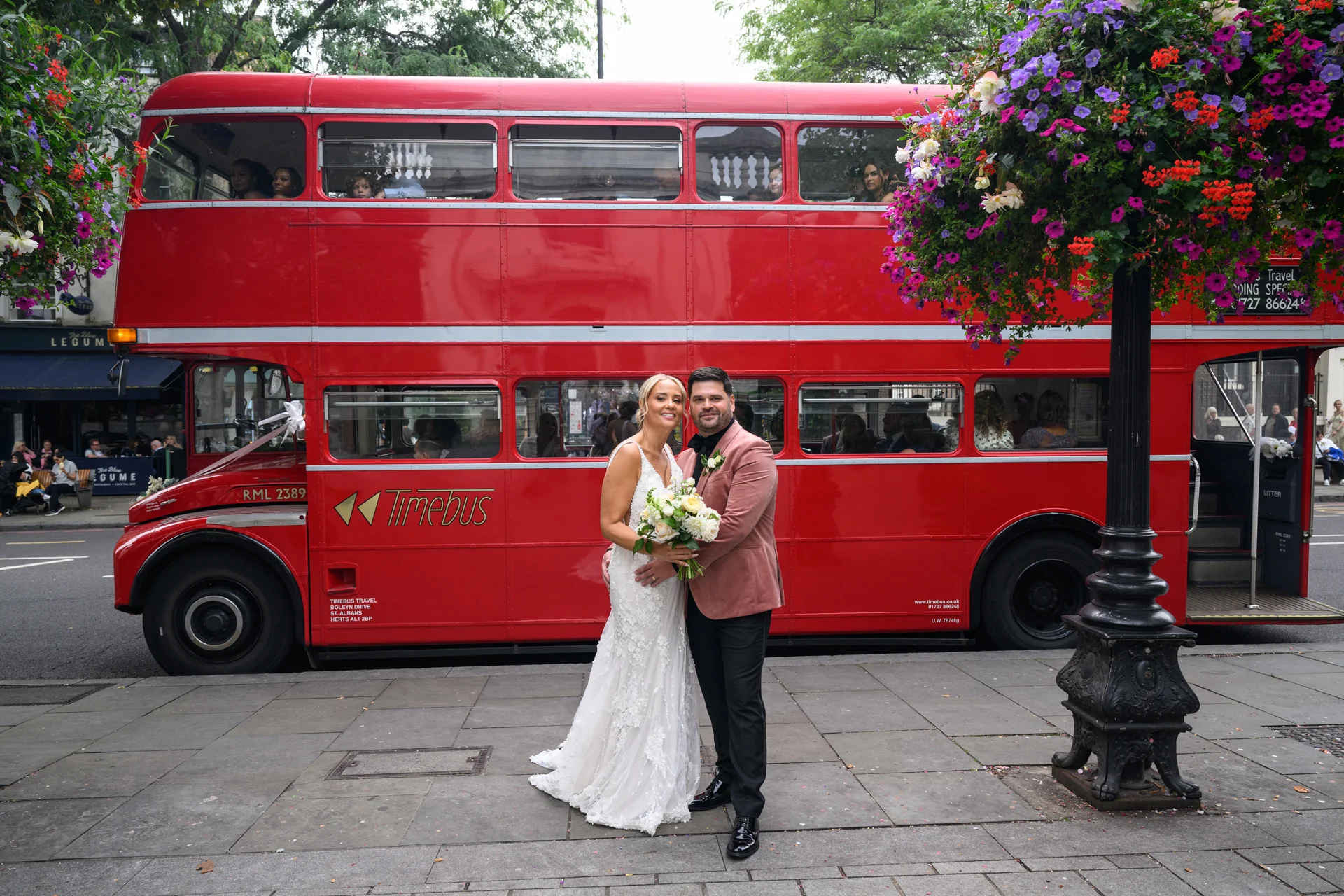 A bride and groom pose happily on a London pavement with a classic red double-decker bus parked in the background.