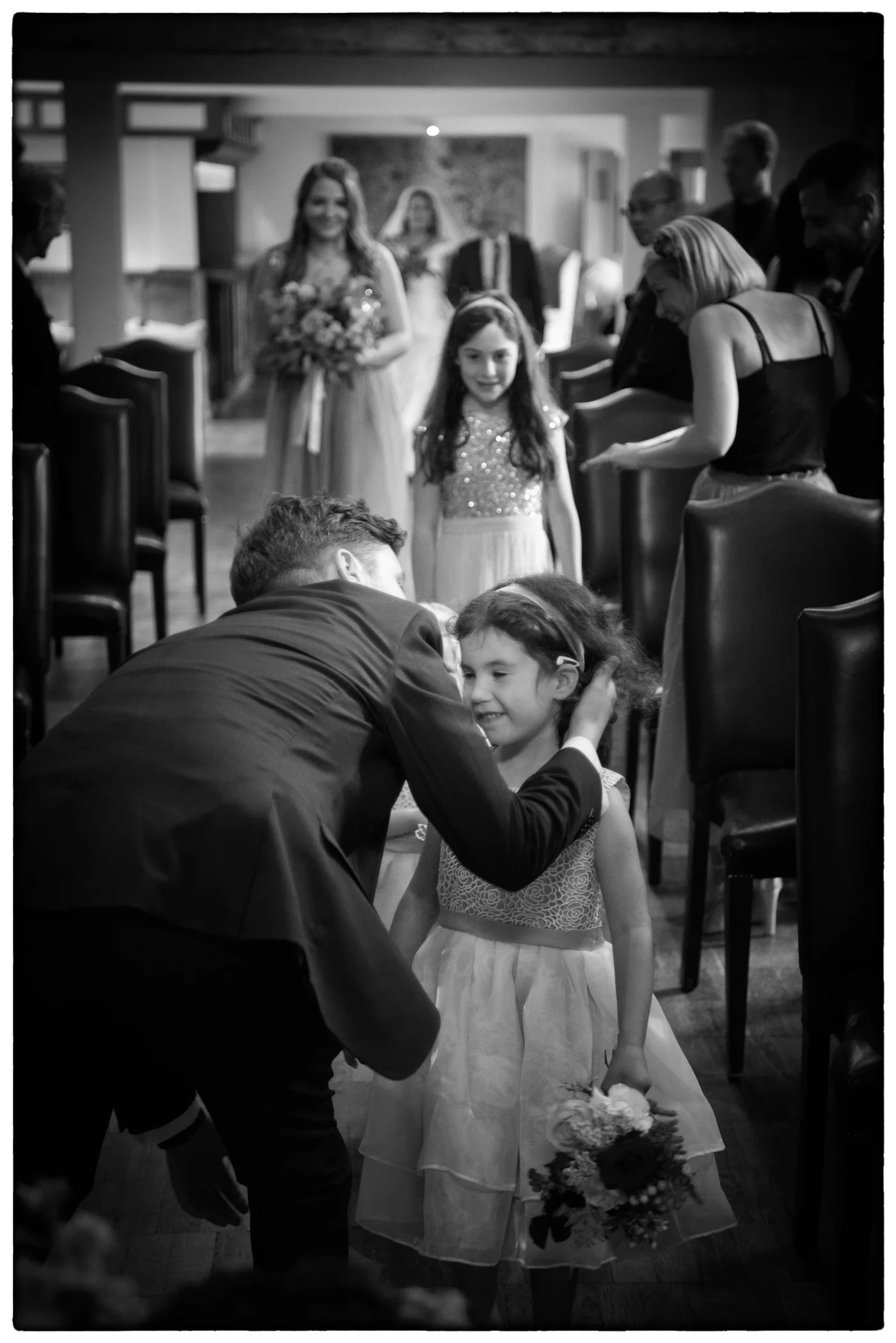 A groom leans down to affectionately greet a smiling young flower girl holding a small bouquet as guests look on during the wedding ceremony aisle processional.
