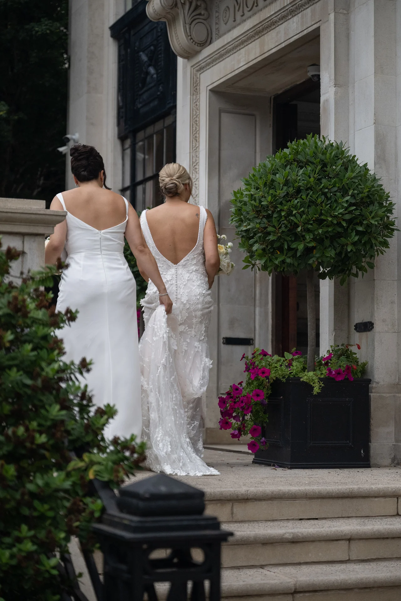 Two brides in white wedding dresses walk hand in hand up stone steps toward a grand hotel entrance.