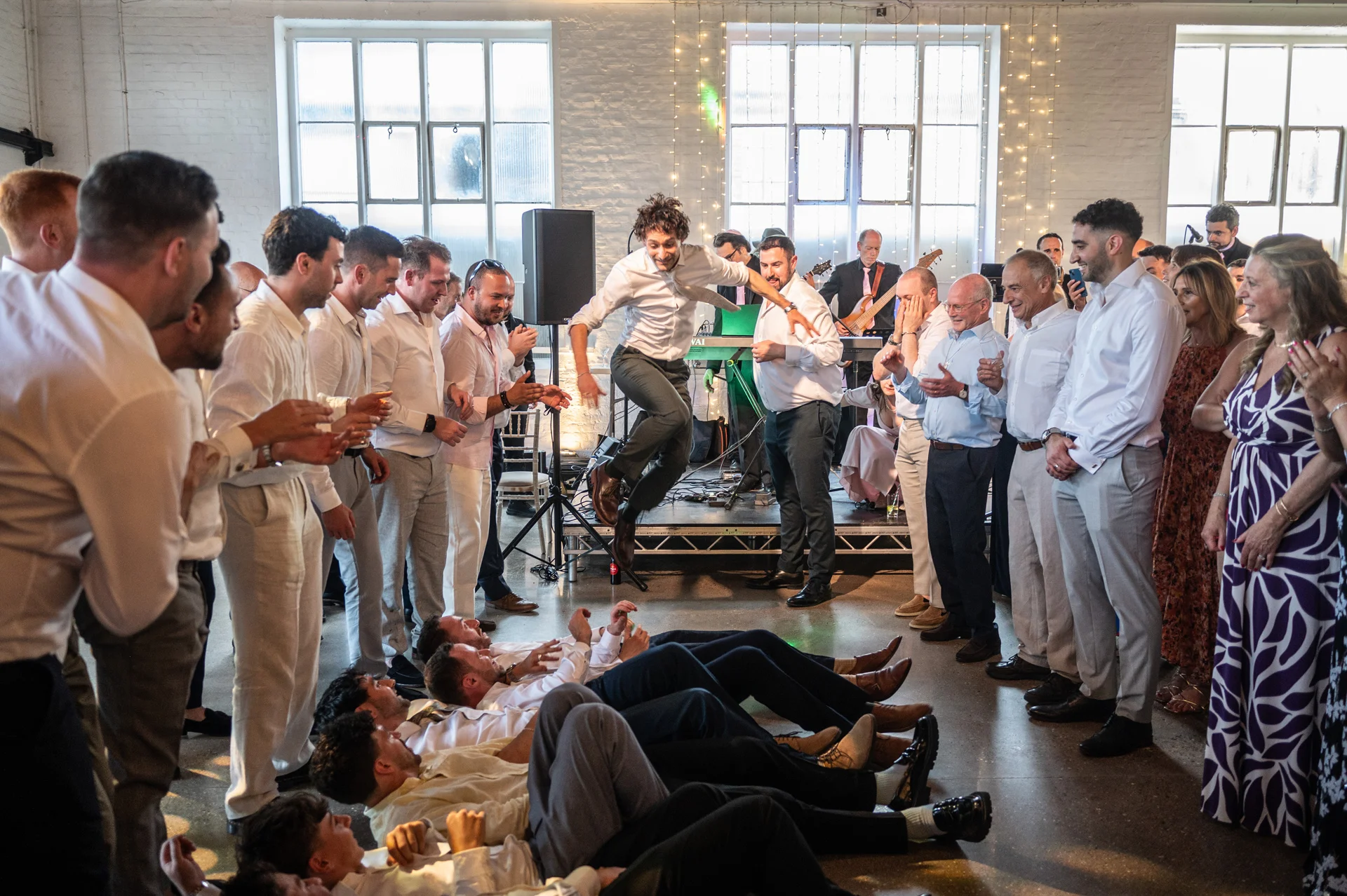 A groom is lifted high in the air by cheering guests on the dance floor of an industrial warehouse venue while a band plays in the background.
