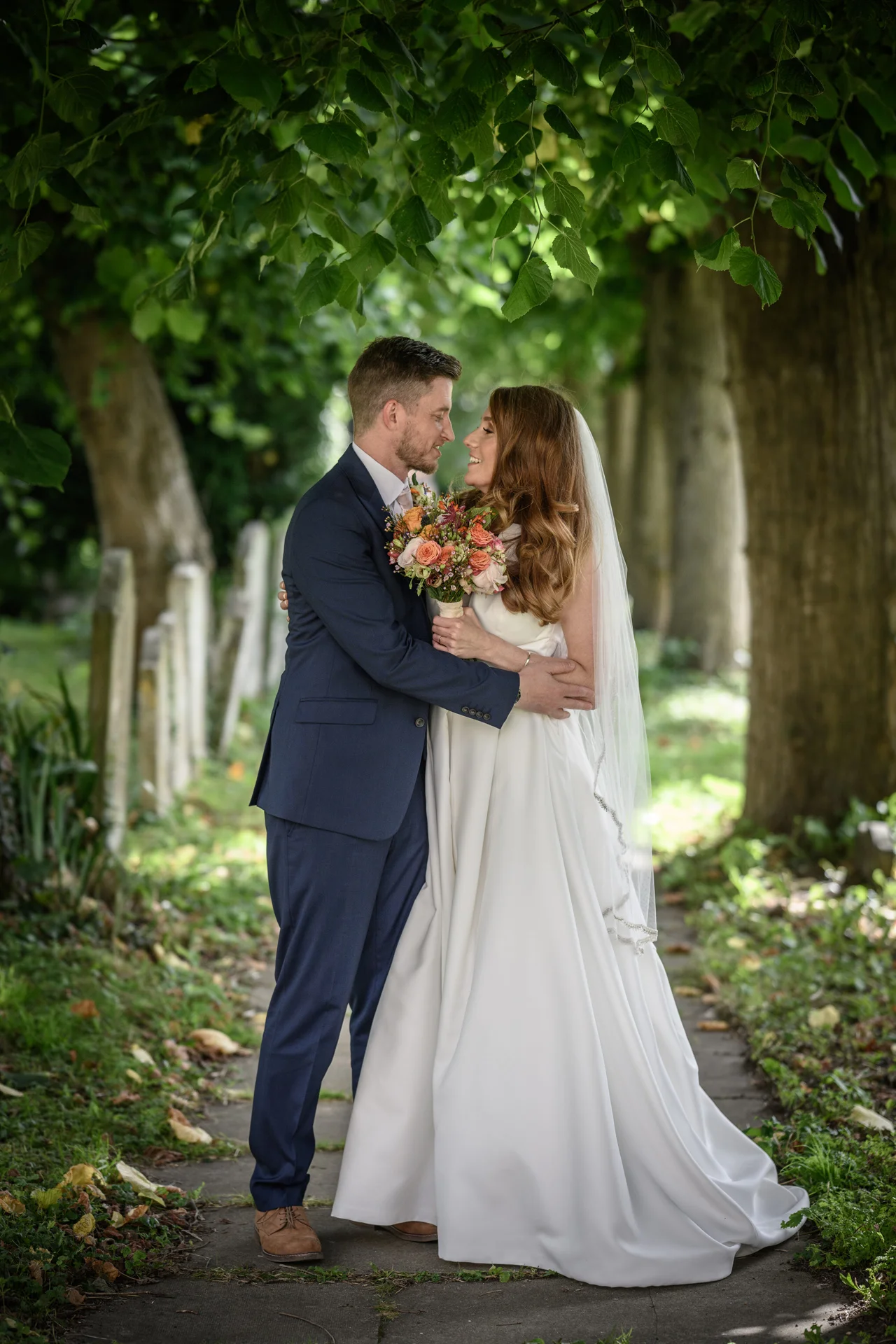 A newlywed couple share a tender nose-to-nose moment while standing along a shaded pathway lined with mature trees.
