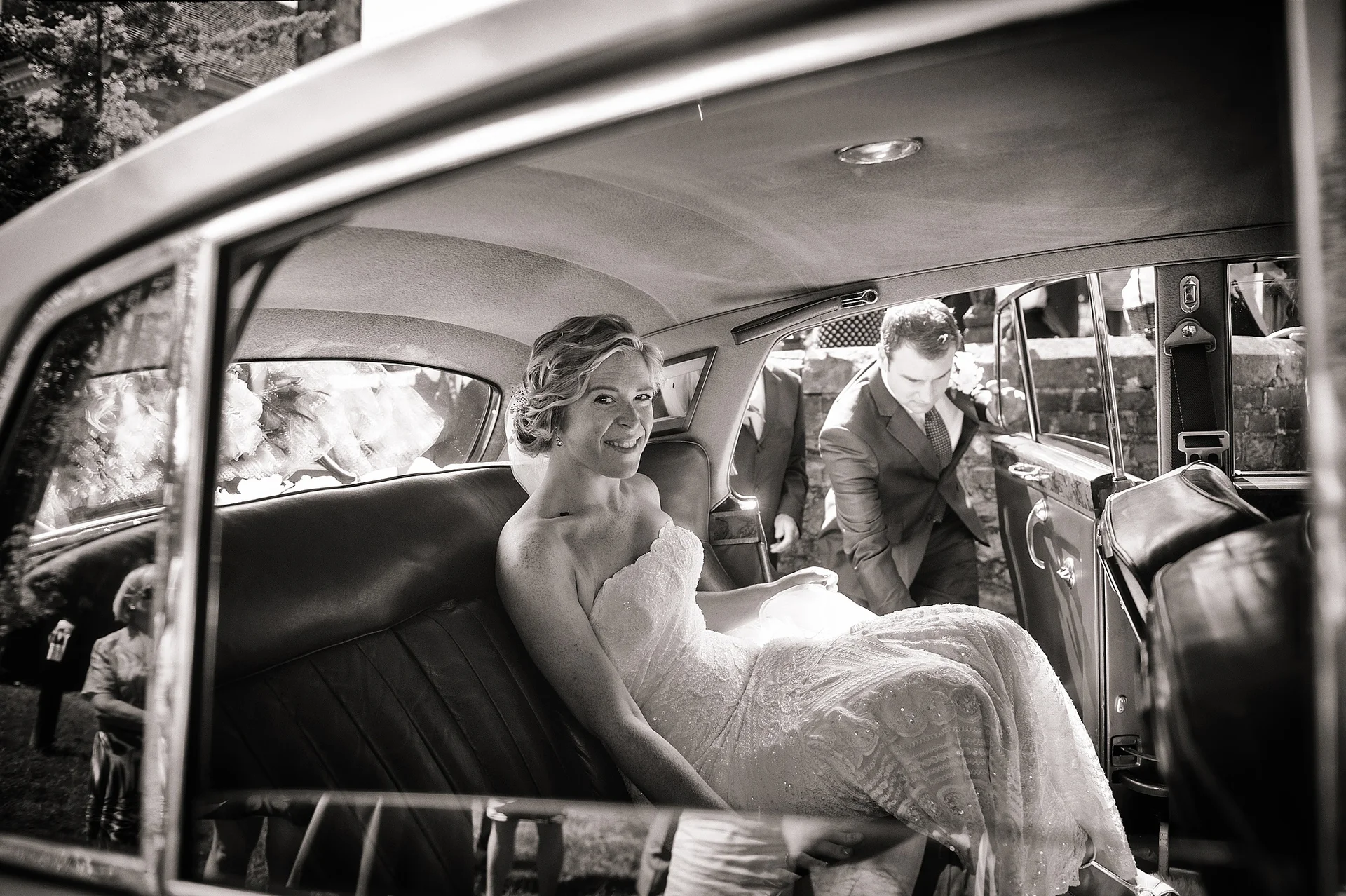 A smiling bride in a strapless lace wedding dress looks out from the back seat of a classic wedding car as she prepares to exit.