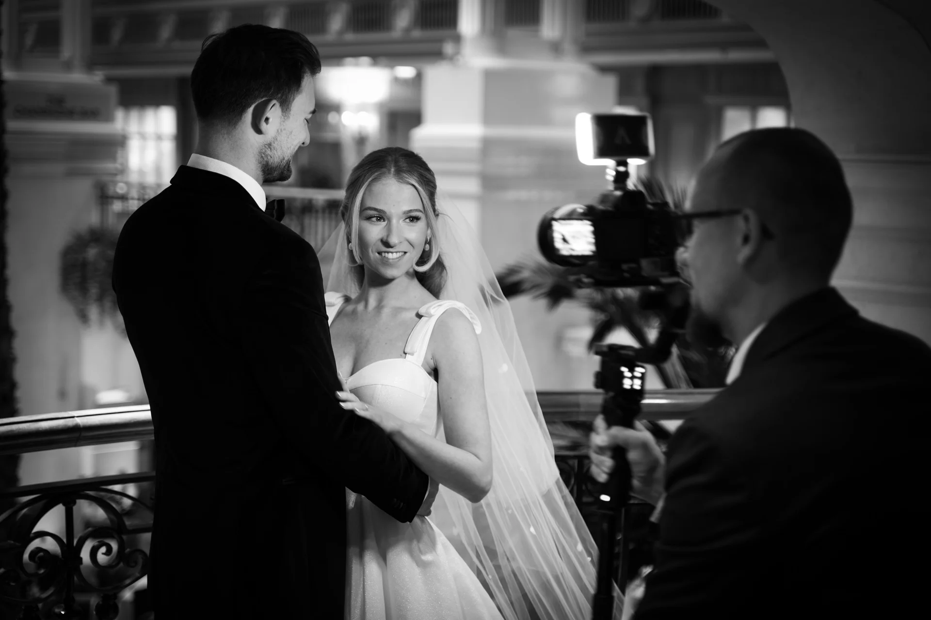 A newlywed couple shares a tender moment on an indoor balcony while being filmed by a videographer in a black and white shot.