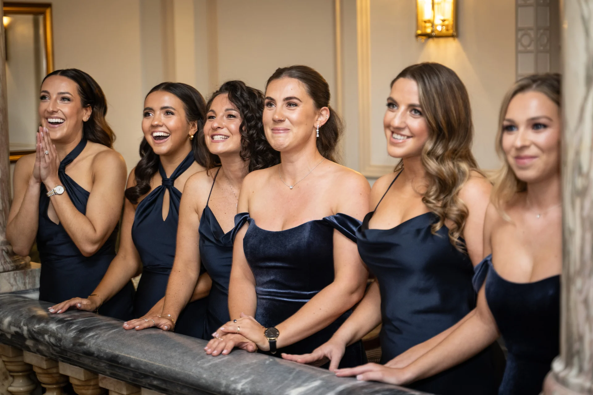 A line of bridesmaids in elegant navy blue dresses smiling and watching joyfully from a grand indoor balcony.