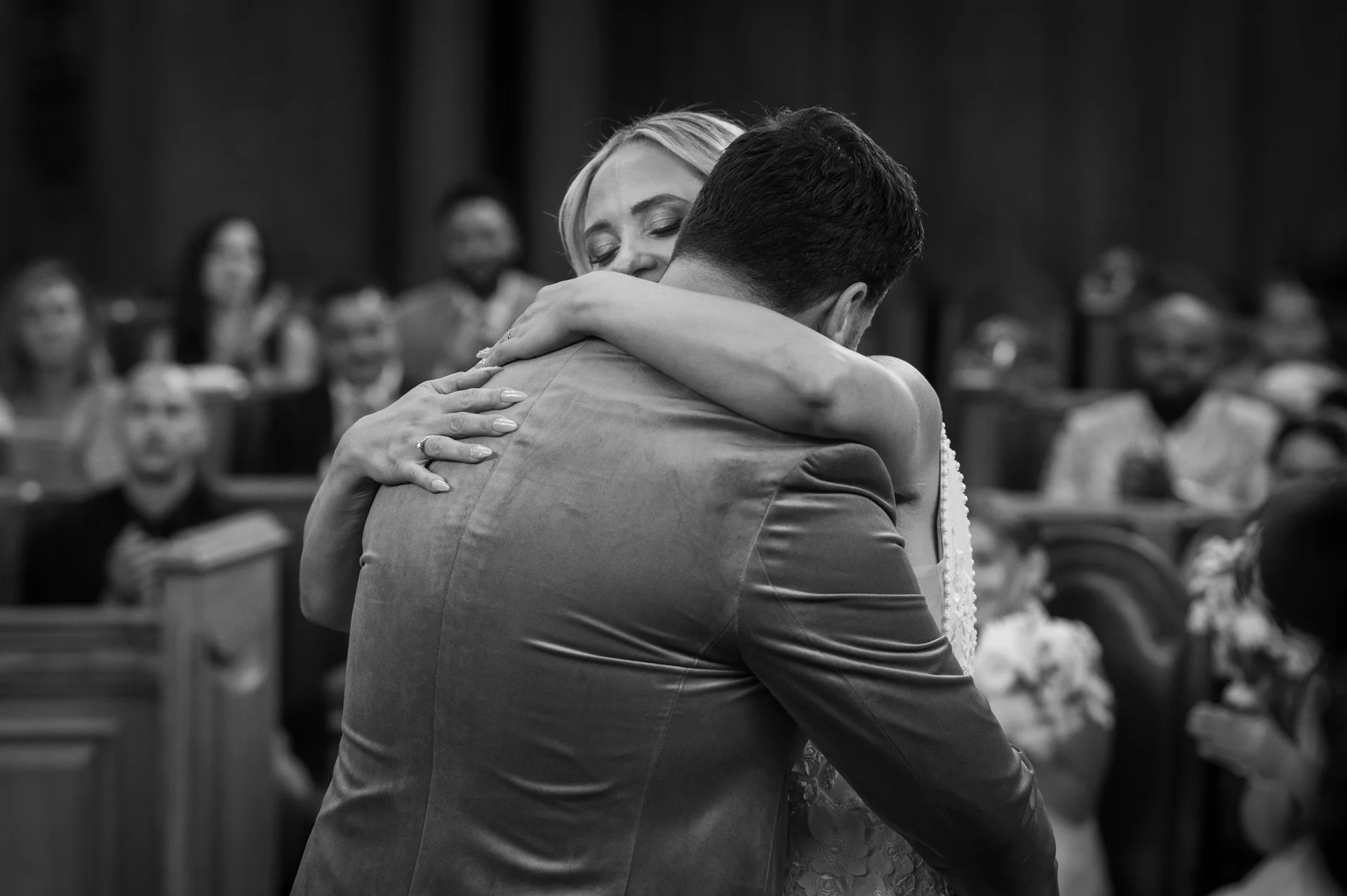 A newlywed couple shares an emotional embrace and a tender laugh during their church wedding ceremony, viewed from over the groom's shoulder.