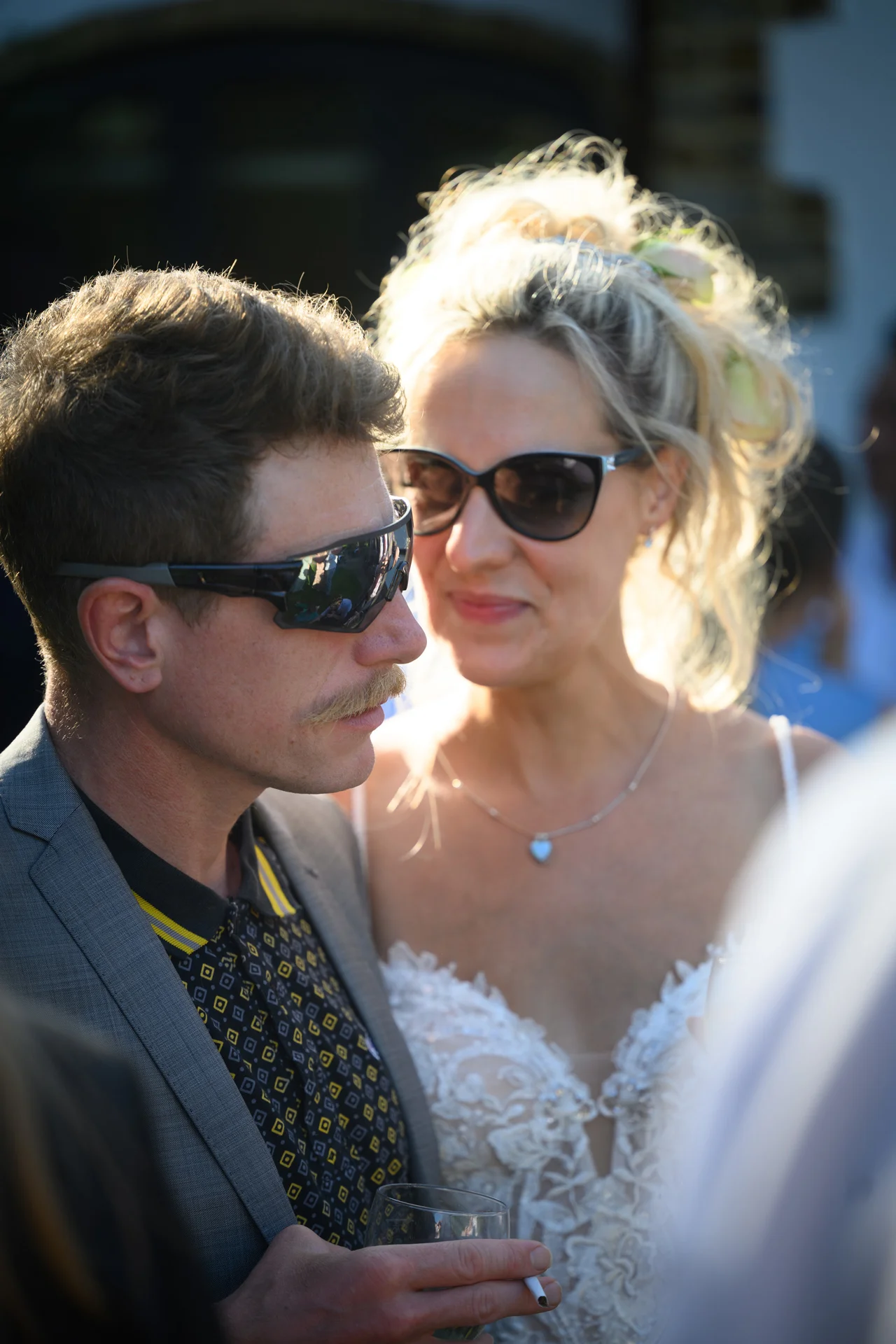 A bride and groom wearing sunglasses share a candid moment outdoors, bathed in warm backlit sunlight.