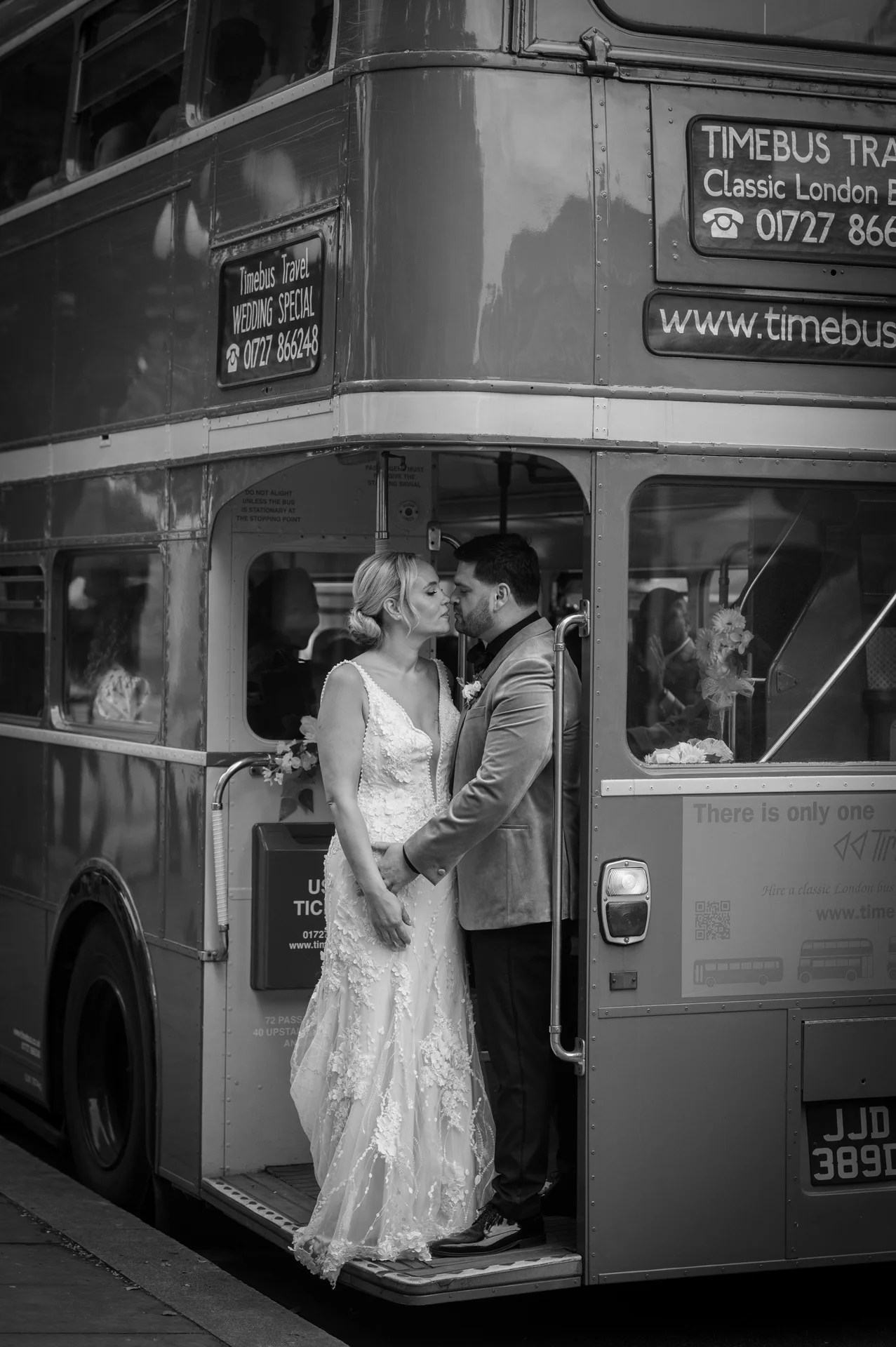 A newlywed couple shares a tender kiss on the steps of a classic red London double-decker bus during their city wedding.