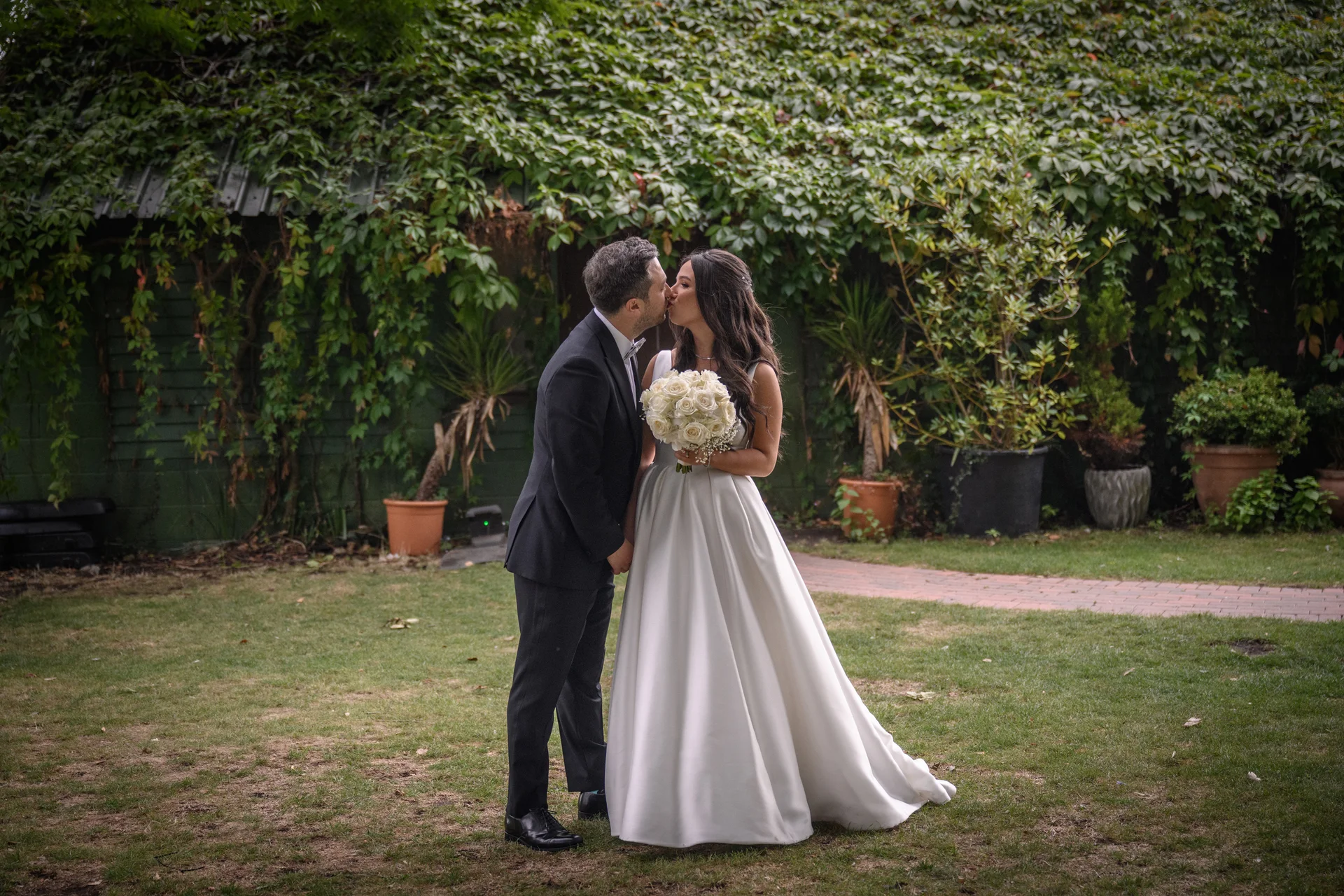 A newlywed couple shares a tender kiss in a lush garden setting with an ivy-covered wall backdrop, the bride holding a classic white rose bouquet.